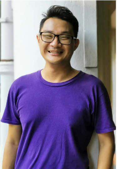 Smiling person wearing a purple T-shirt and glasses, standing indoors against a light-colored wall.