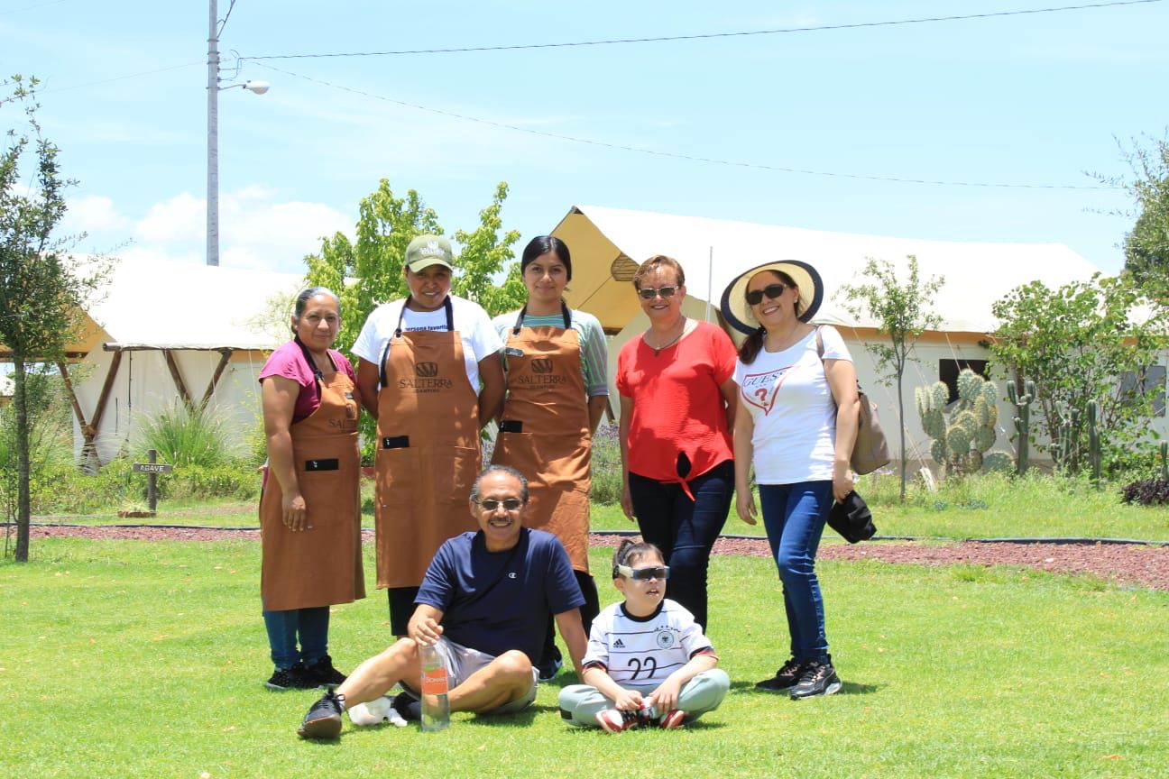 Group of seven people posing outdoors on grass, some wearing aprons. Tents in the background, sunny day with trees nearby.