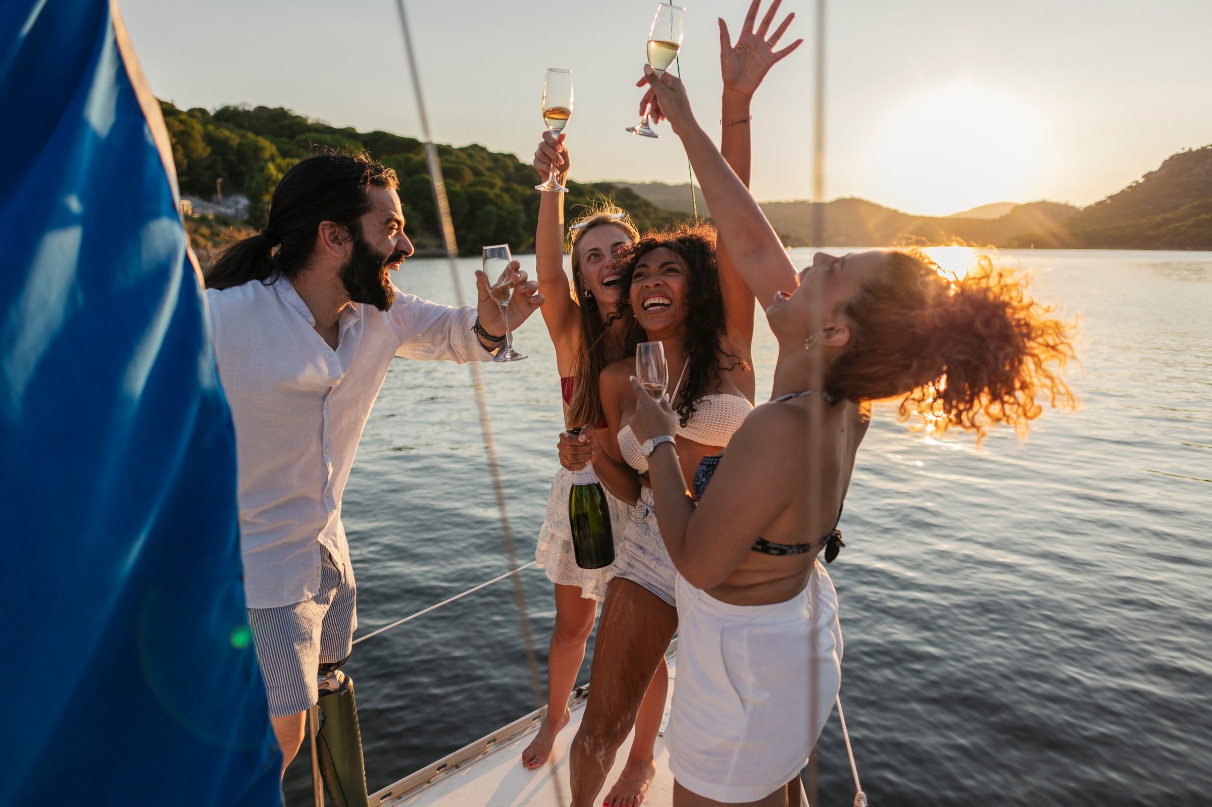 Young friends are cheering and raising champagne glasses on a yacht during a golden sunset, enjoying a summer vacation and celebrating friendship together on the open water