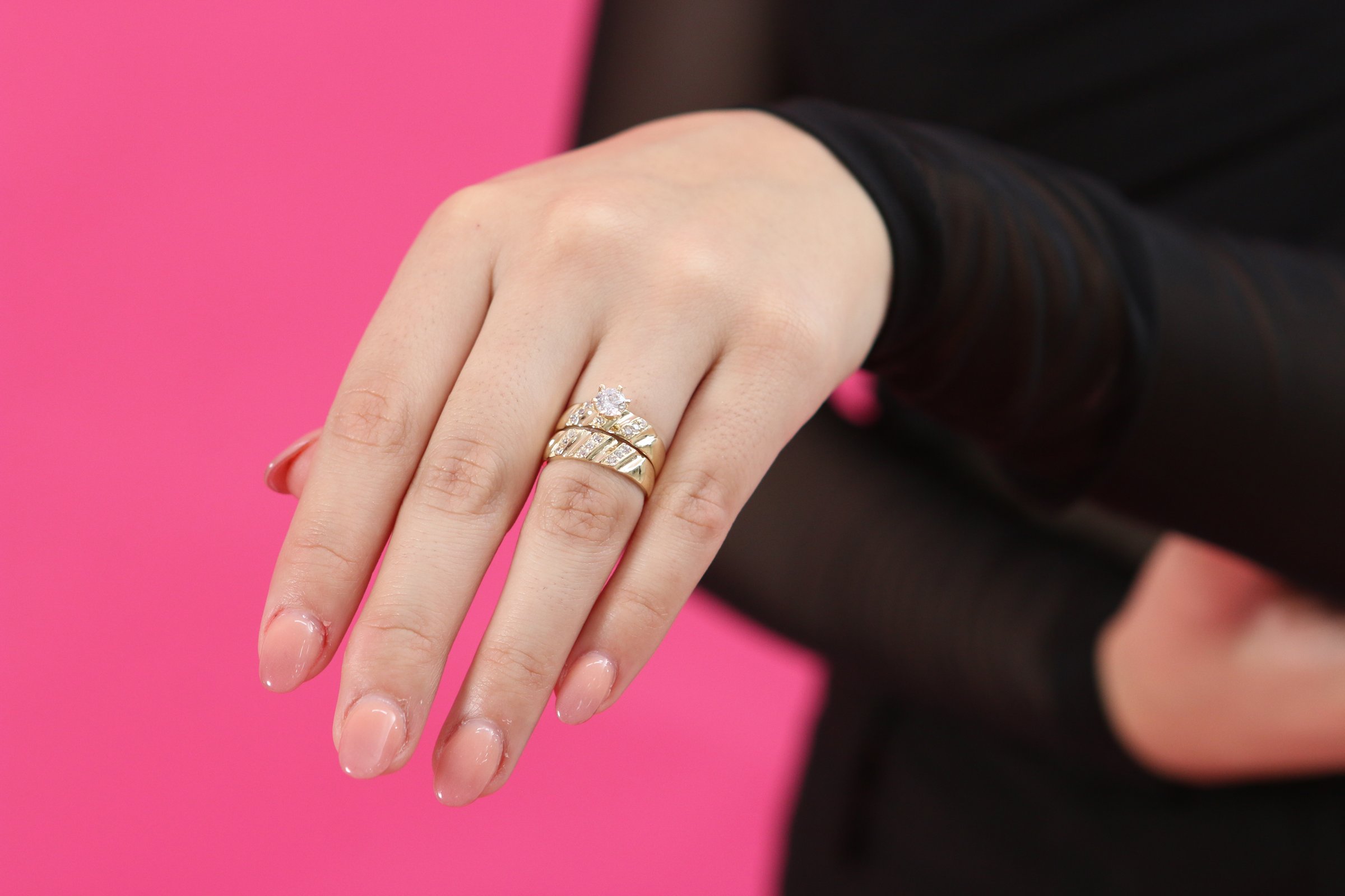 A hand with manicured nails wears a gold ring with a diamond, against a pink background.