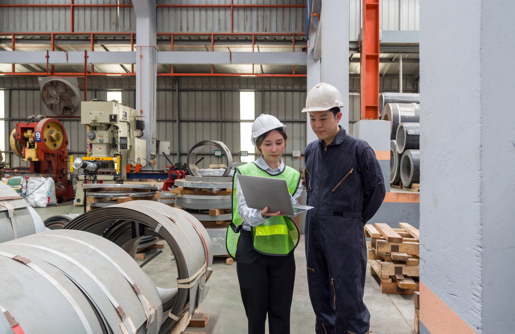 Two people are working in a factory. They are discussing plans while looking at a laptop. They wear safety helmets.