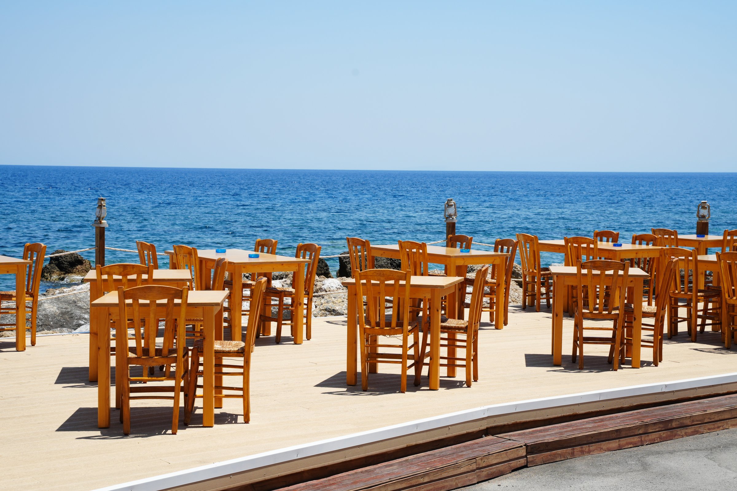 Empty outdoor restaurant terrace with wooden tables and chairs overlooking a calm ocean. The setup is perfect for alfresco dining, offering a serene and inviting atmosphere.