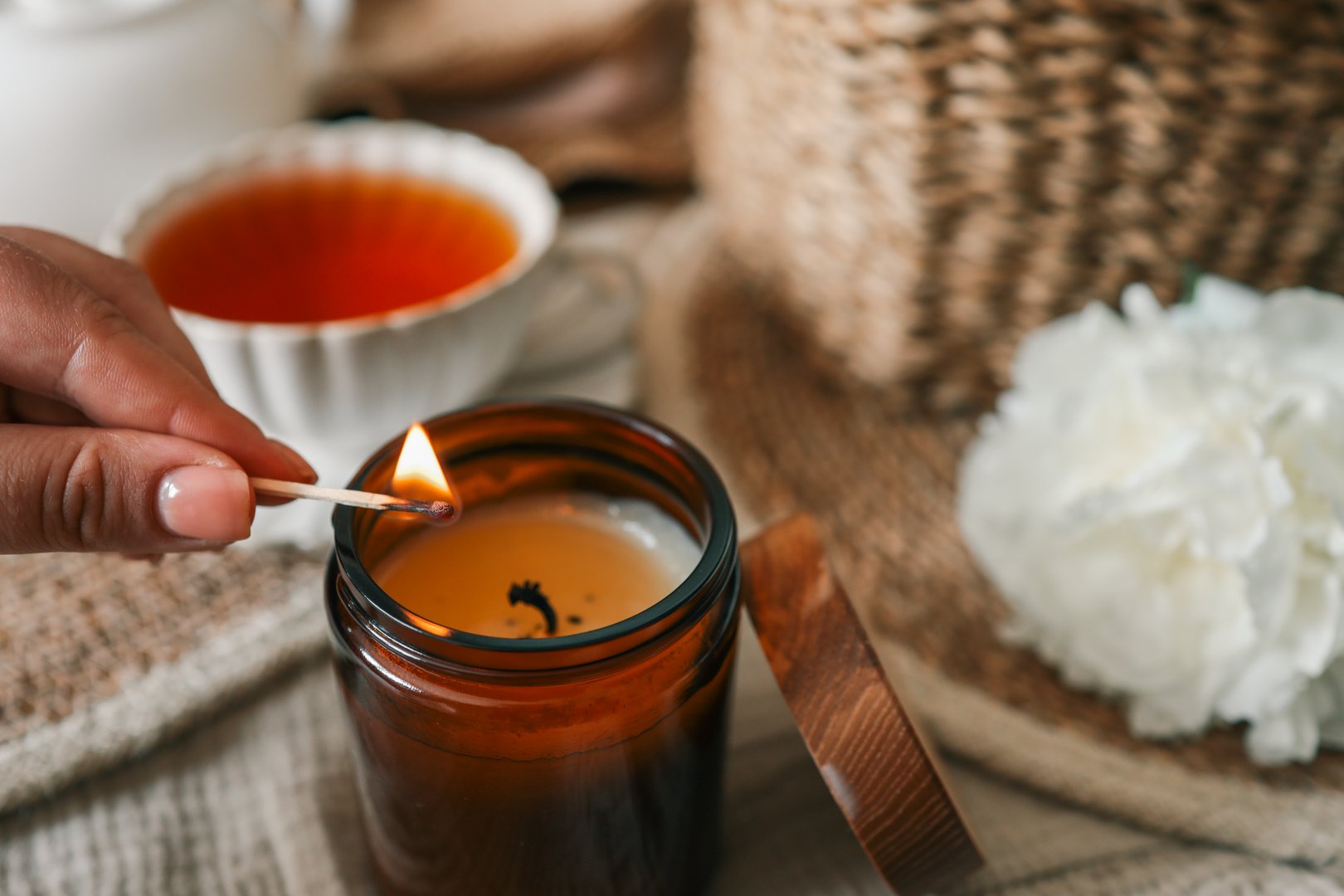 Hand lighting a candle in a cozy flat lay scene with tea, white peonies, straw hat, and an open book. Warm summer moment of relaxation and self-care in soft natural light.