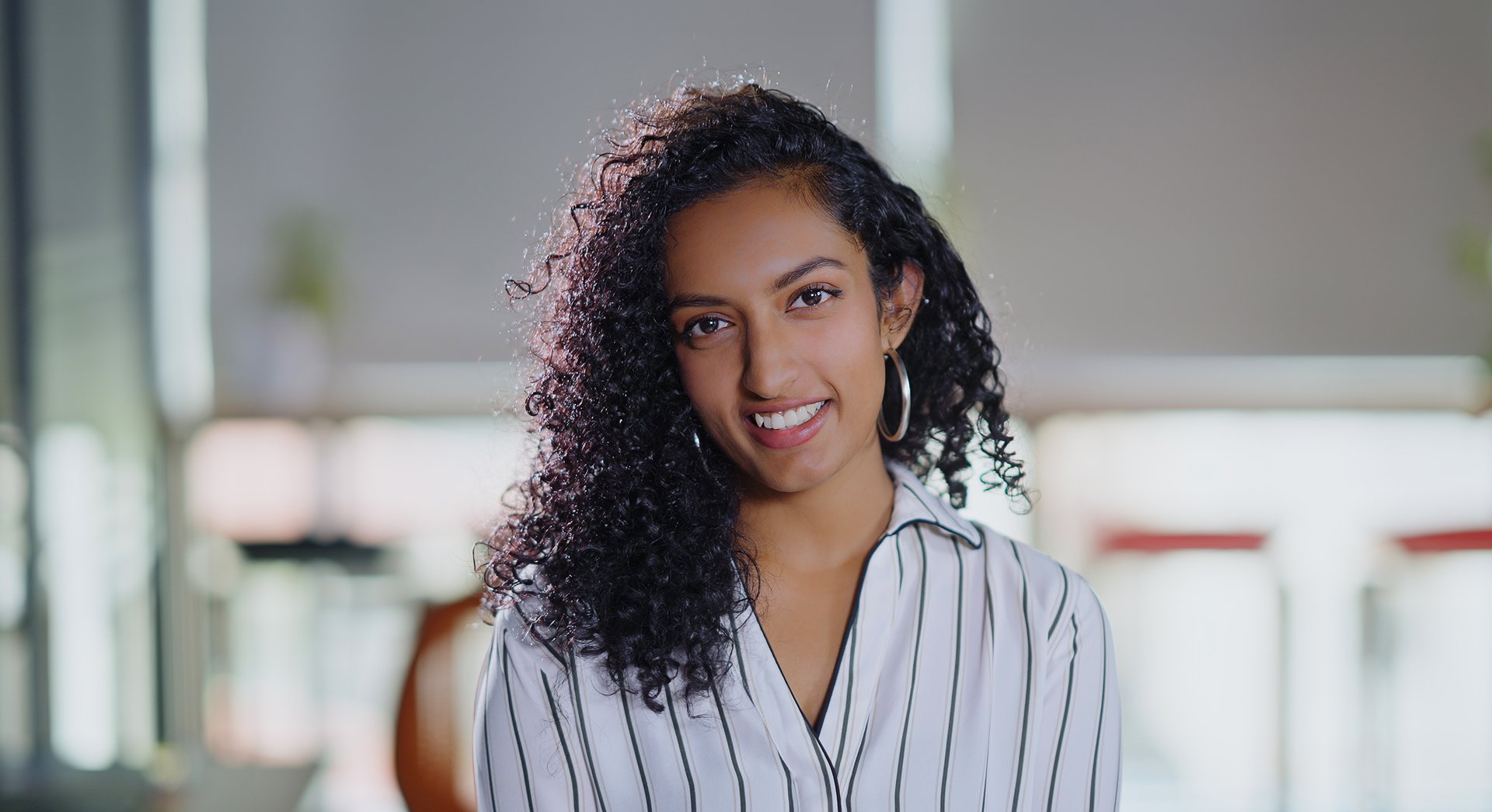 Indian curly hair young adult gen z business woman lady expert intern staff worker standing proud look cam indoor office place. Confident happy smiling cute teen corporate girl do pose head shot portrait photo at job workplace