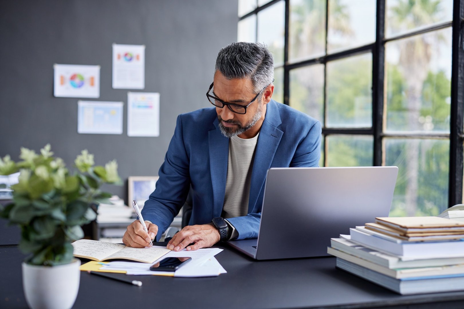Middle eastern business man in blue blazer working at desk with laptop while taking notes. Mid adult professional man writing in notebook in office. Thoughtful smart casual businessman reviewing documents in modern workspace with copy space.