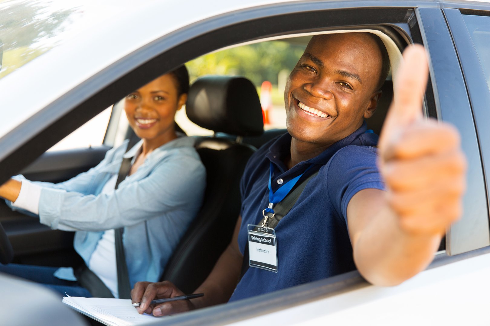 happy male african driving instructor in a car with learner driver giving thumb up