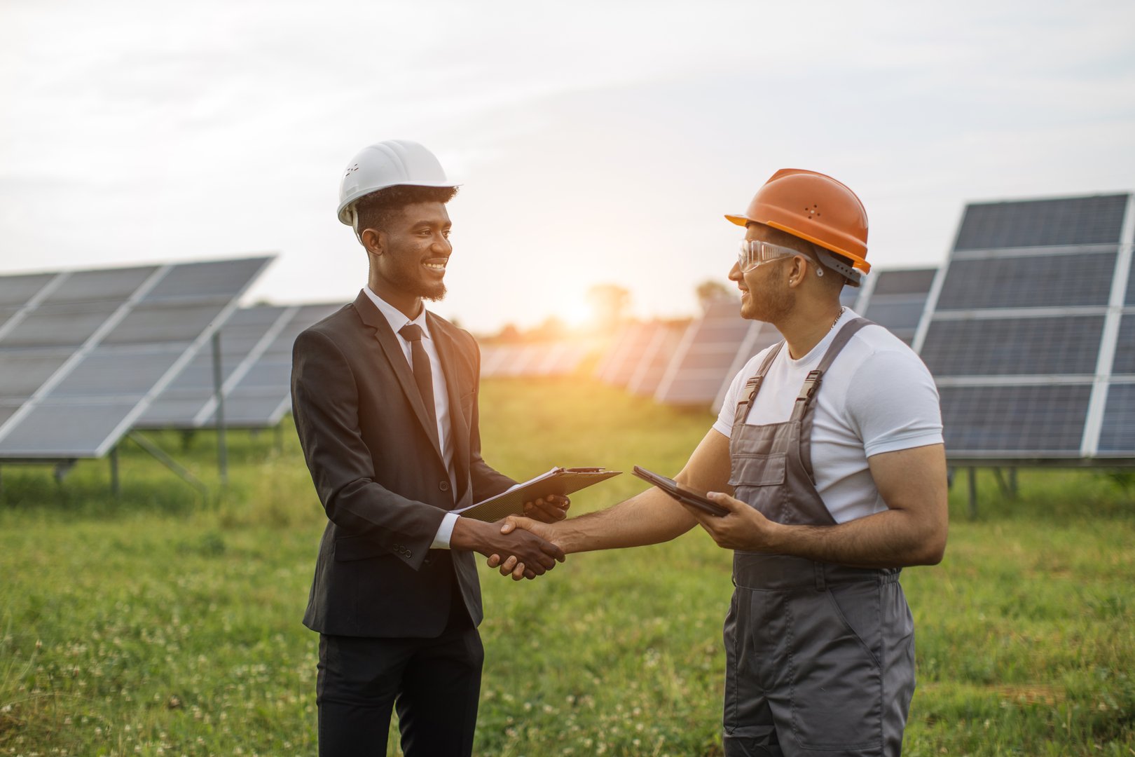 African american man in suit and white helmet shaking hands with indian man in uniform and yellow hard hat. Inspector and engineer standing together among solar plant.