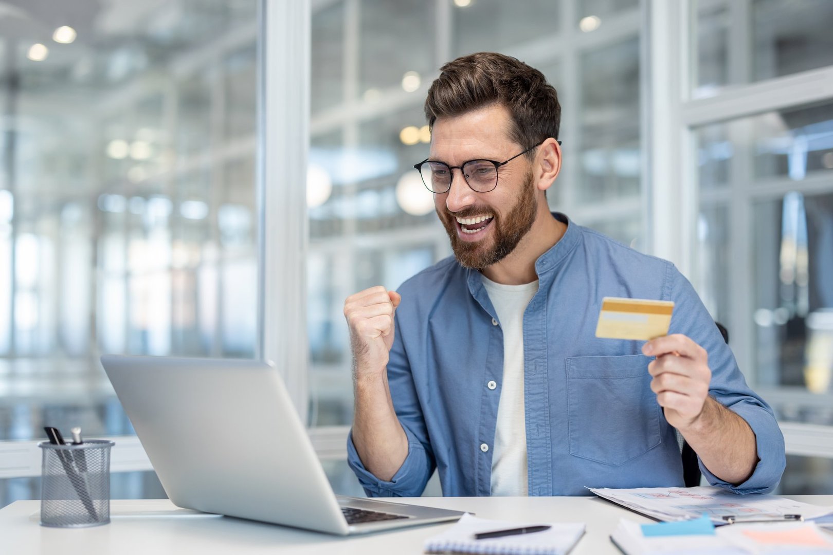 A man celebrates a successful online purchase, holding a credit card near a laptop in an office setting. He is joyful.