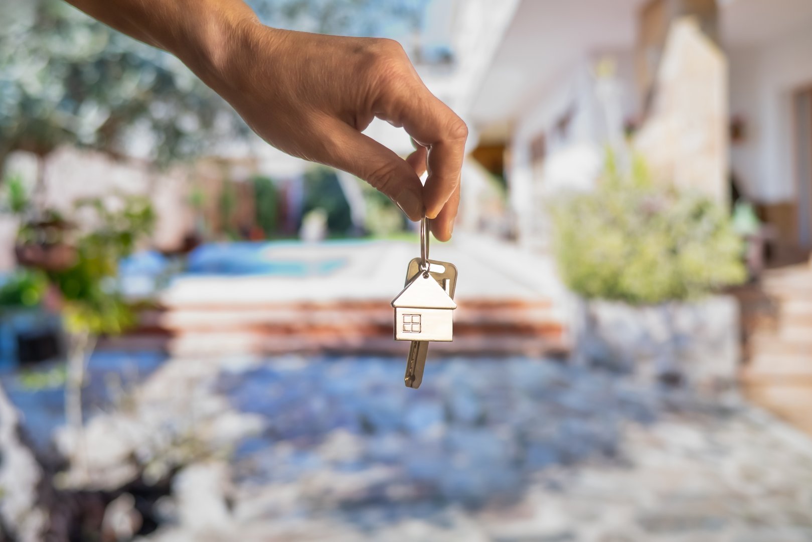 Hand holding house keys with house shaped keychain in front of a blurred background representing a new home with a swimming pool, symbolizing real estate investment and homeownership