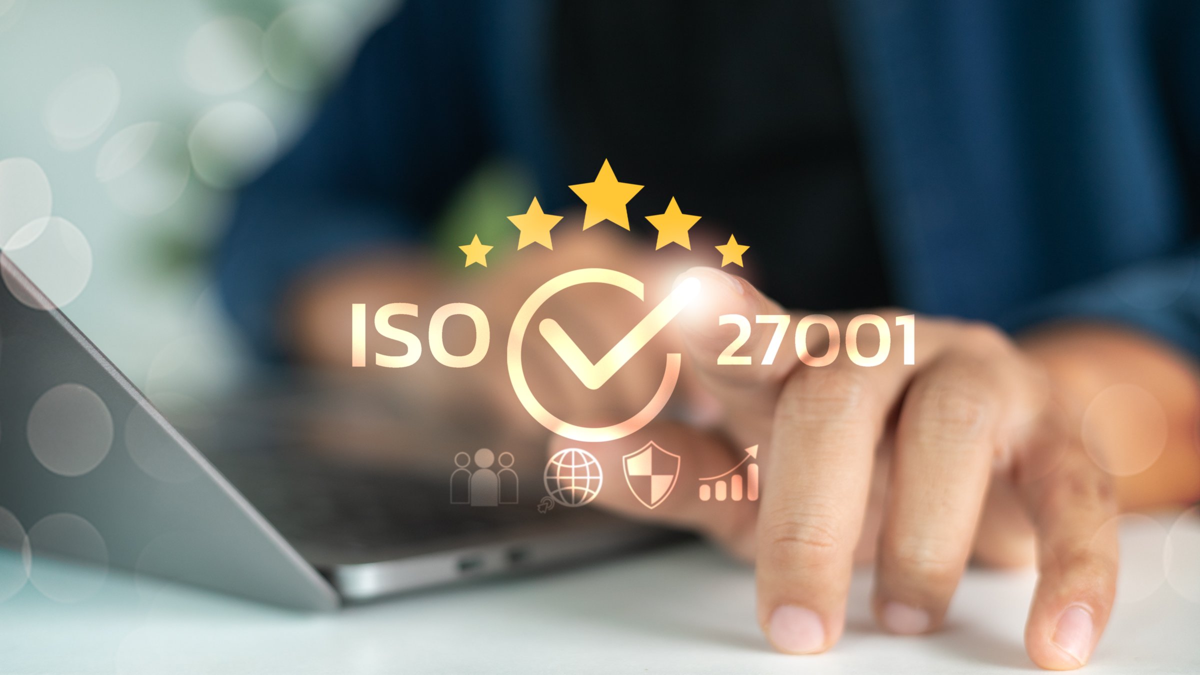 Close-up of a hand using a laptop with ISO 27001 certification icon, check mark, and five stars, representing cybersecurity and data protection standards.