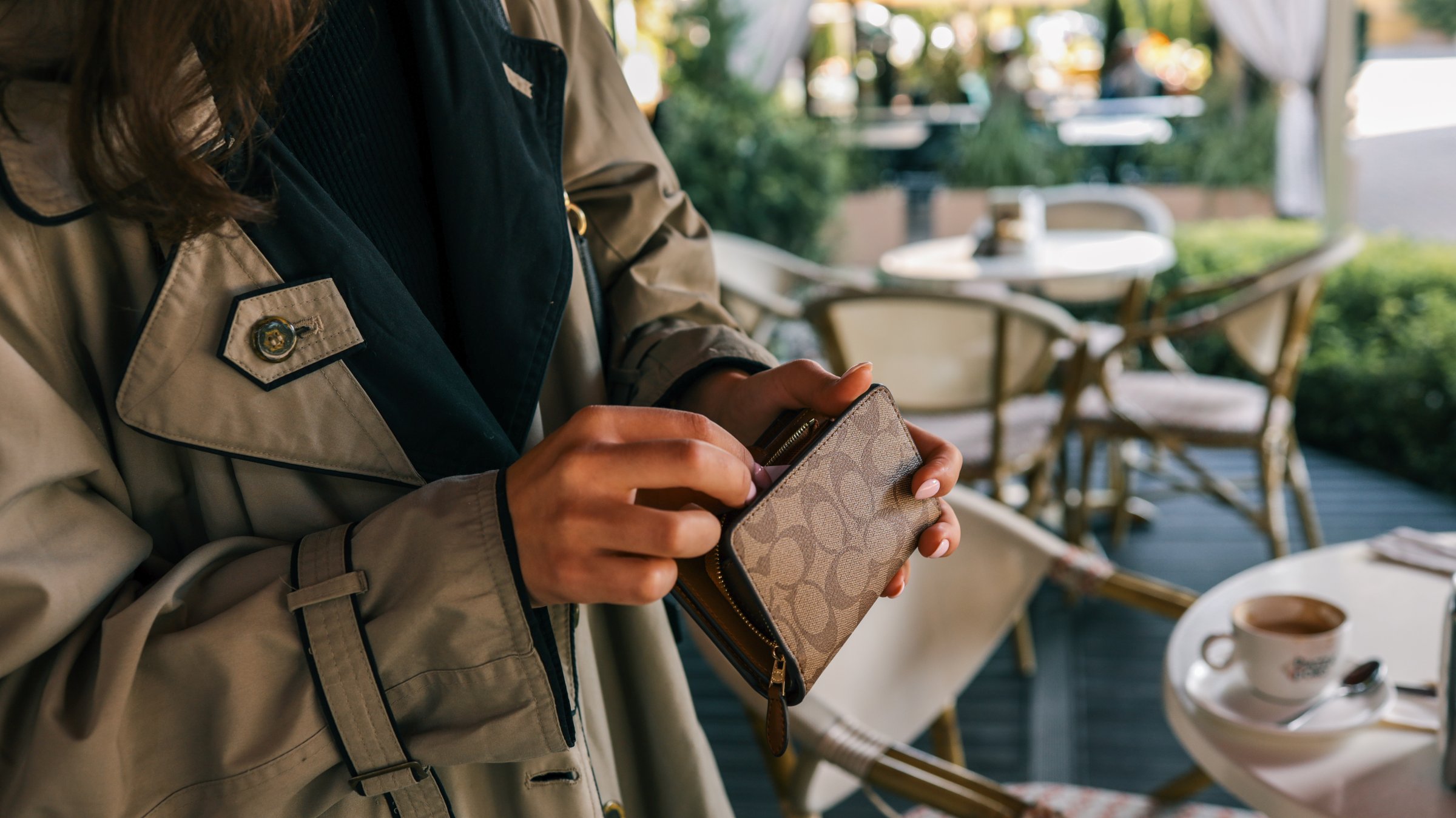 Close-up of woman's hands in beige trench coat unzipping a stylish brown wallet with monogram pattern, with blurred cafe background and coffee cup on table.