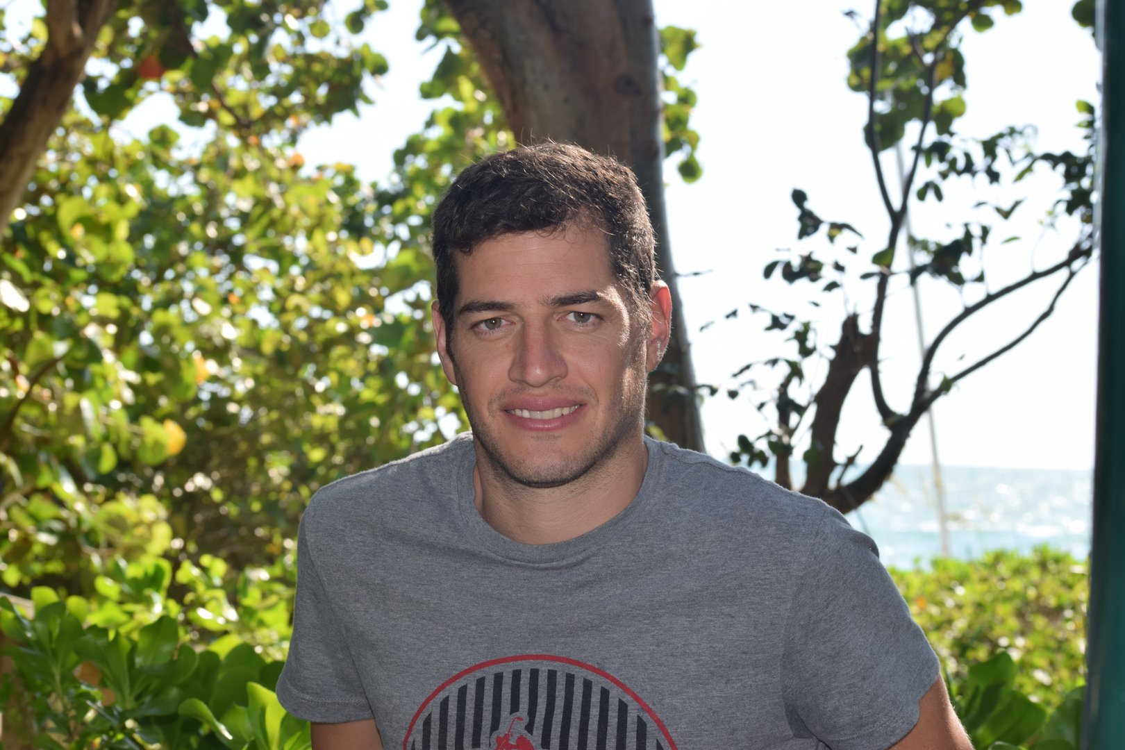 A man in a gray shirt stands outdoors, surrounded by lush greenery and trees, with the ocean visible in the background.