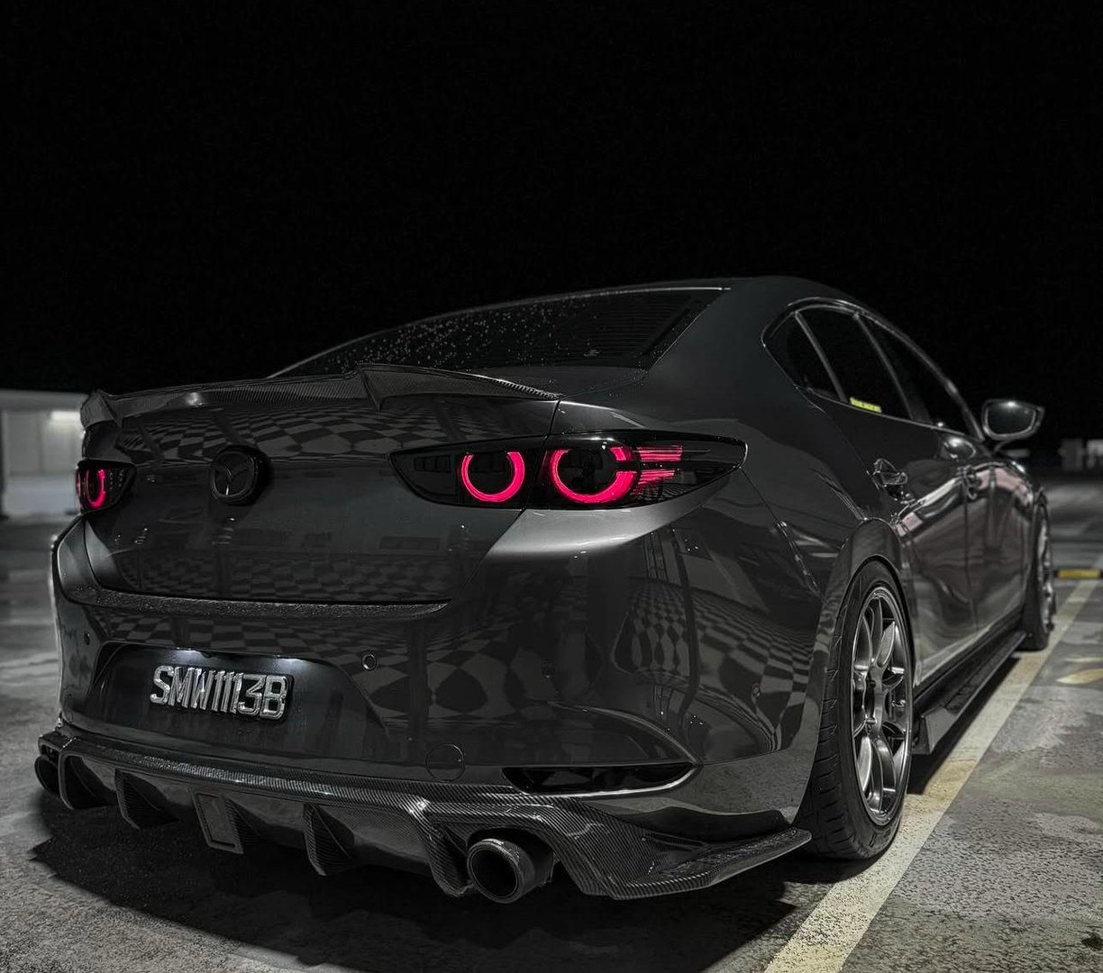Modified black car at night with glowing red taillights and a noticeable rear diffuser, parked in an empty lot.