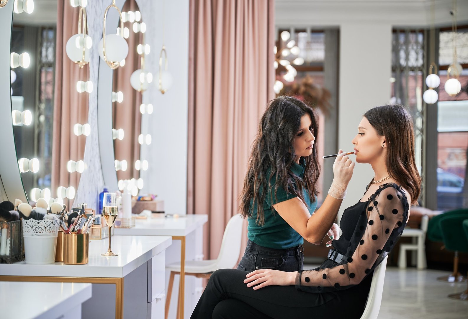 Makeup artist doing professional makeup in visage studio. Stylish woman sitting at dressing table while female beauty specialist in sterile gloves applying lipstick on client lips with cosmetic brush.