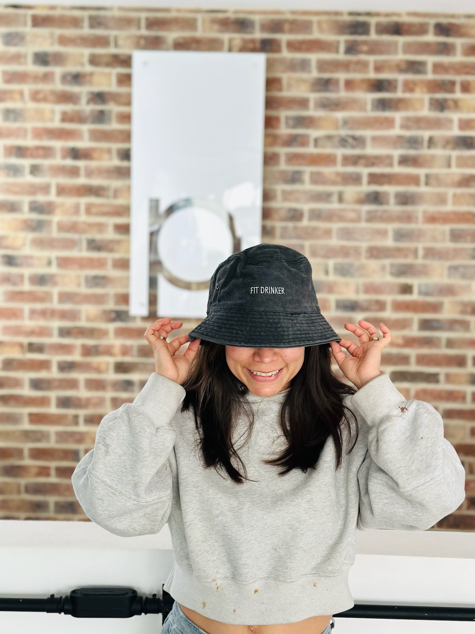Woman in gray sweater and bucket hat smiling in front of a brick wall, partially covering her face with the hat.
