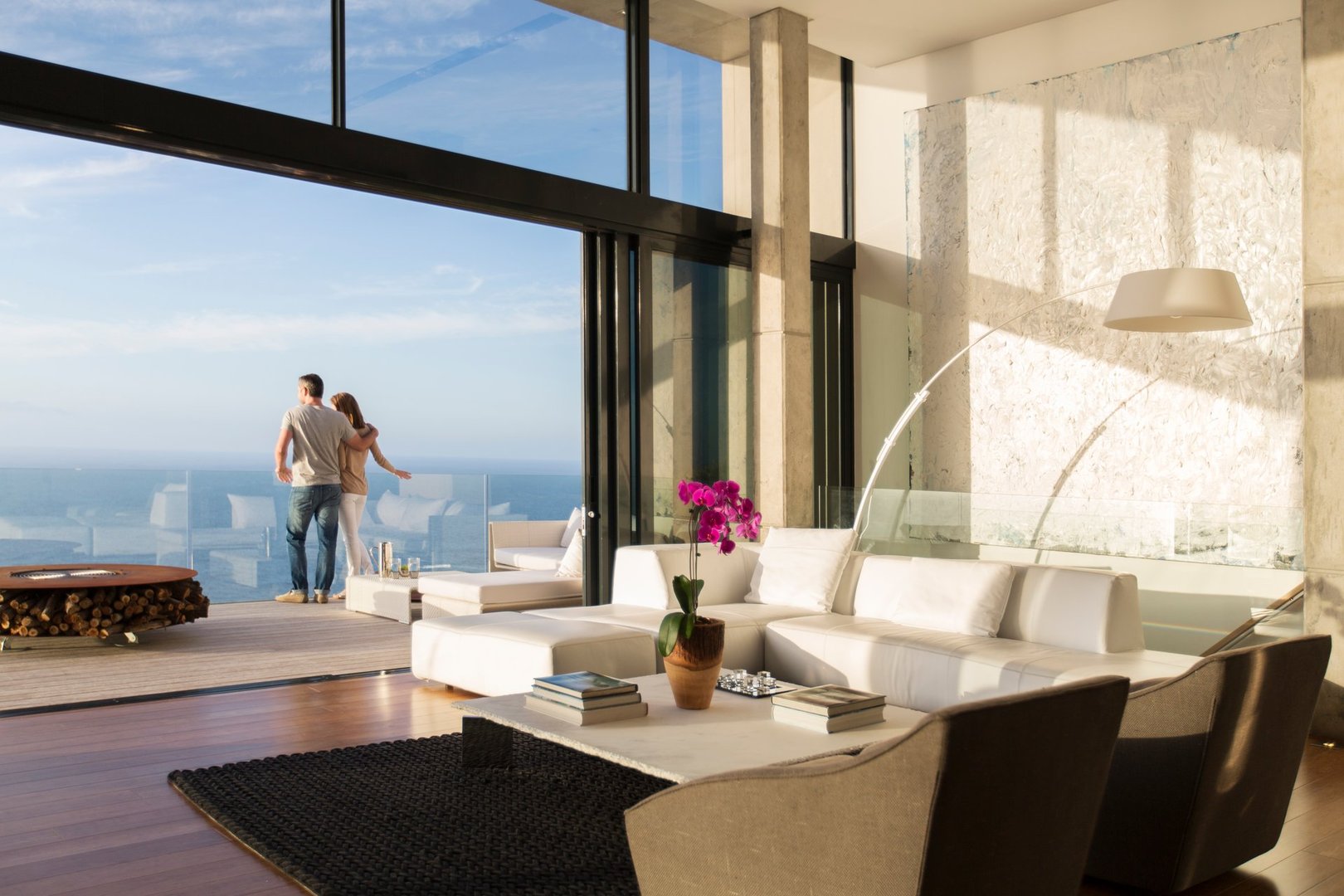 Modern living room with ocean view, white sectional sofa, large windows, and a couple standing on the balcony.