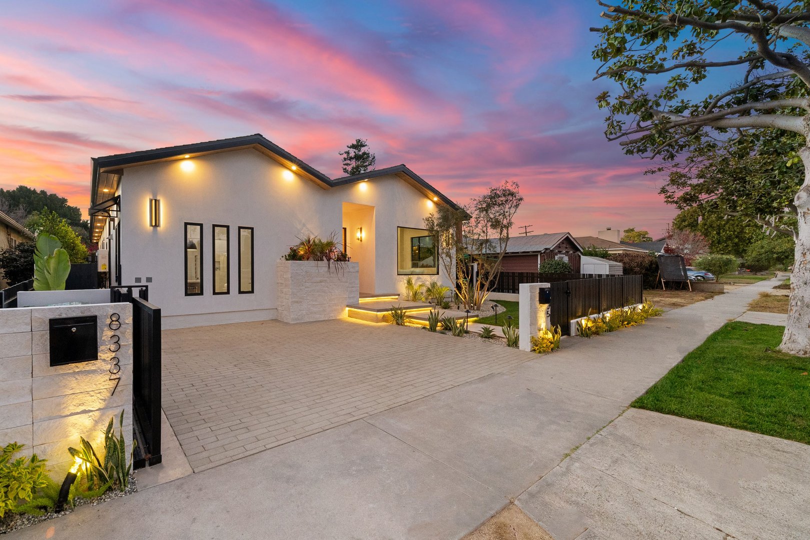 Modern house exterior with illuminated facade and landscaped front yard at sunset, showcasing a vibrant sky.