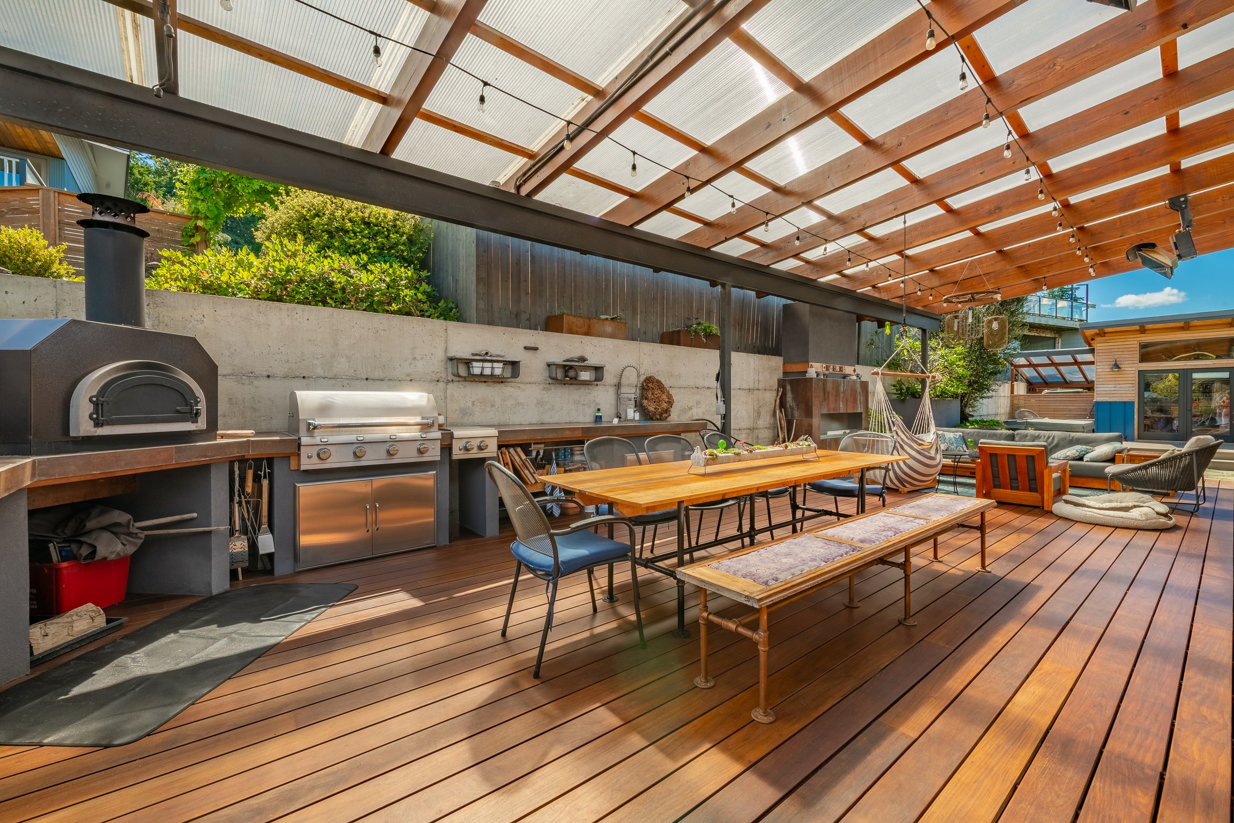 Image displays a large modern outdoor living space, specifically a covered patio and deck area. The space features extensive wooden decking and a slatted wooden roof with translucent panels for light and to shield from rain.