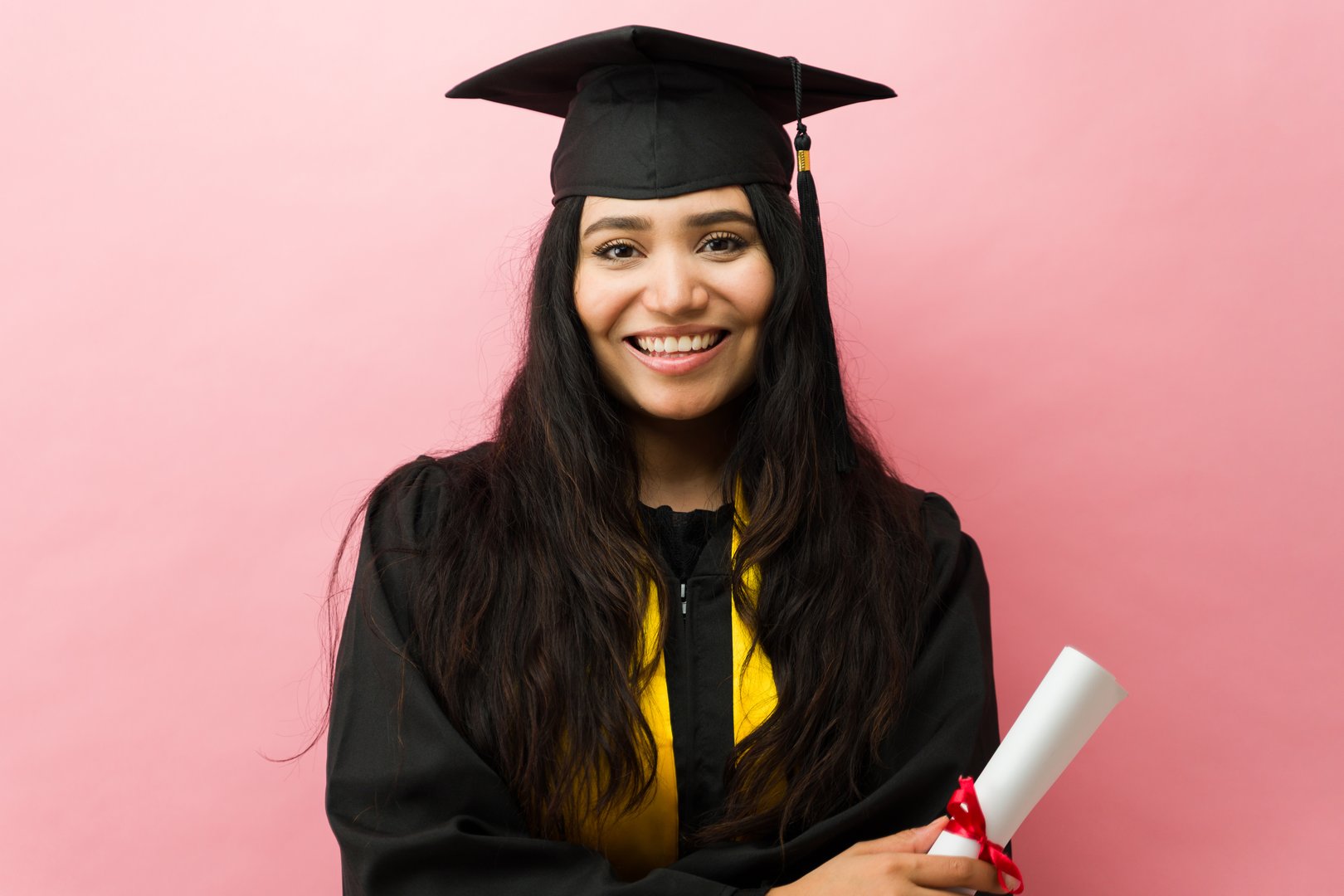 Smiling young woman holding her diploma after completing her studies, wearing a graduation cap and gown in a studio