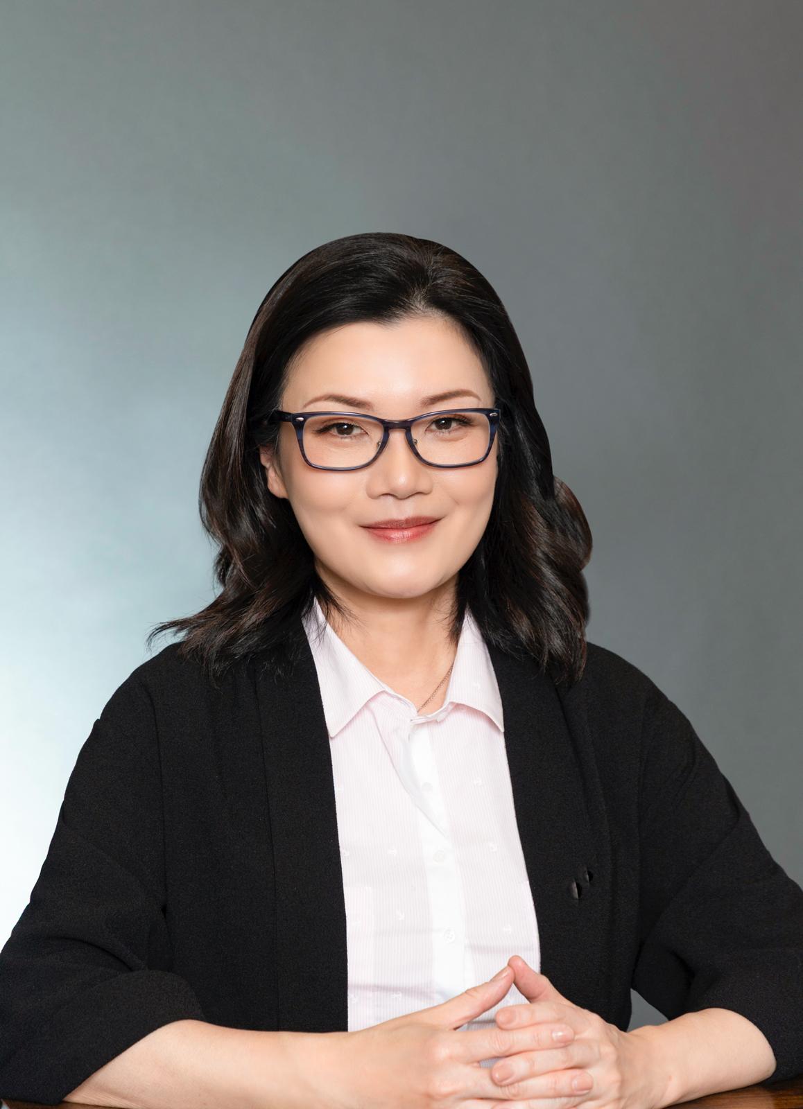Woman with glasses and black hair, wearing a white shirt and black cardigan, smiles with hands clasped.