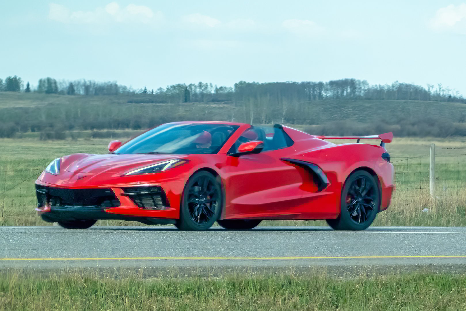 Calgary, Alberta, Canada. May 9, 2025. A vibrant red Chevrolet Corvette Stingray convertible speeds along a rural highway under a bright sky, showcasing its sleek design and performance.