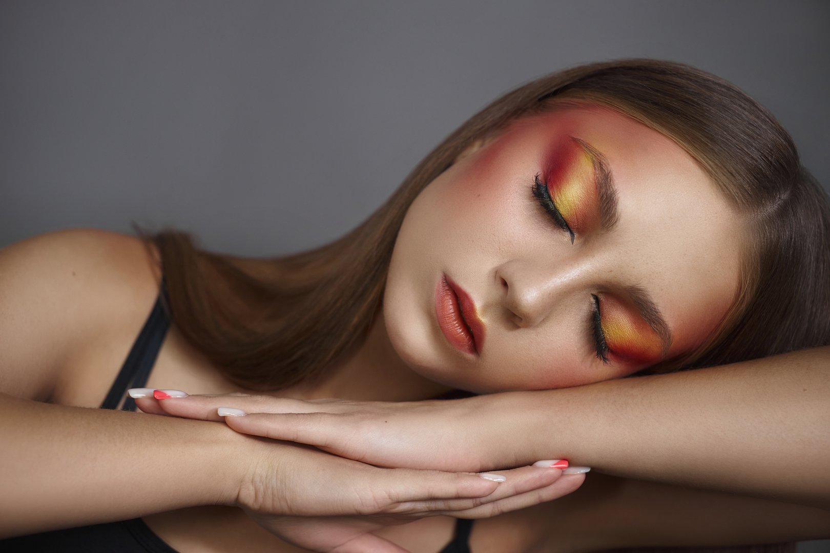 model with long hair rests her head on her arms, showcasing striking warm-toned makeup with orange and yellow hues, creating a captivating artistic expression.