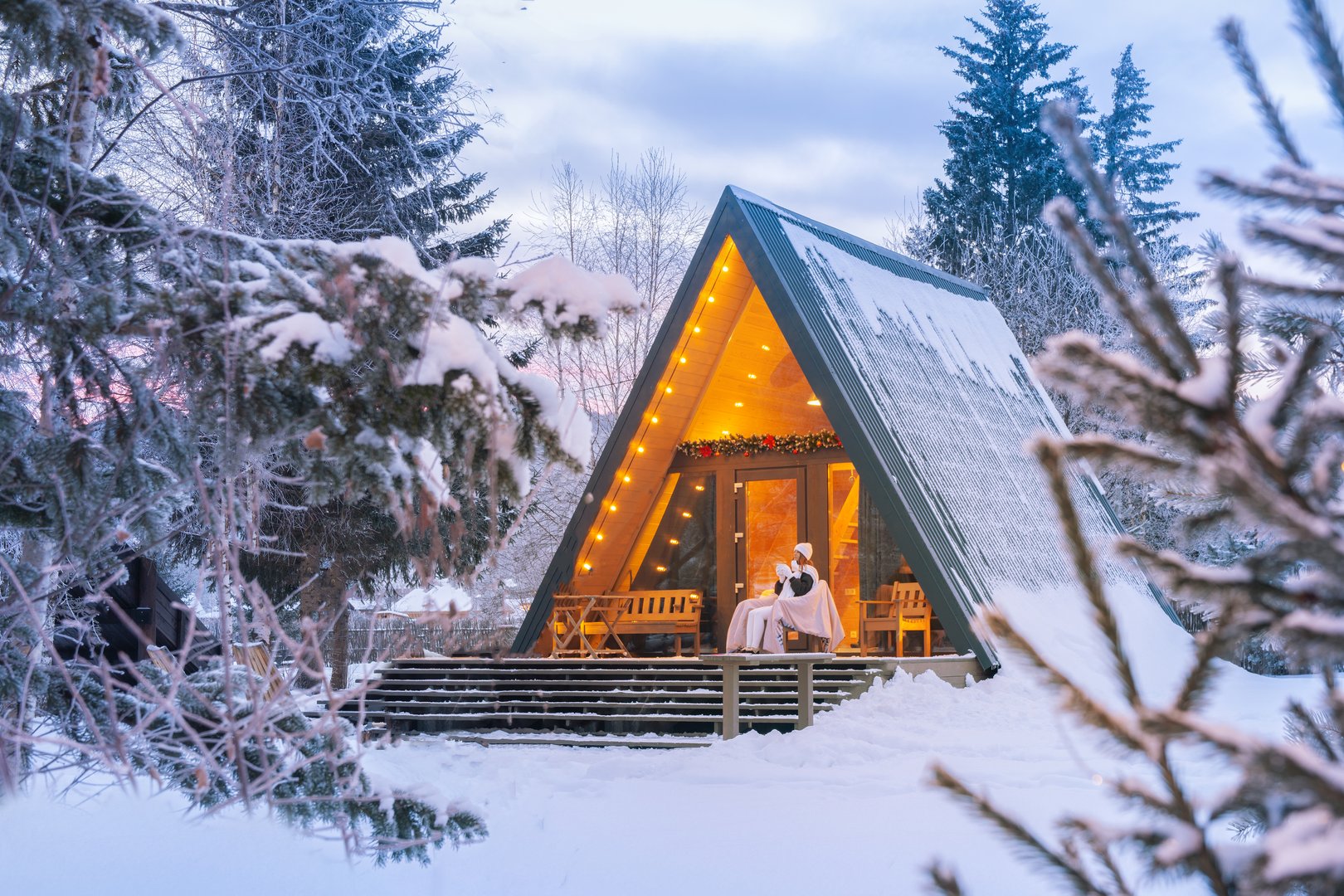 warm drink on the porch of an A-frame cabin in a serene winter forest during the early morning light. A woman drinks coffee or tea in a quiet evening in a house near the forest in the late evening