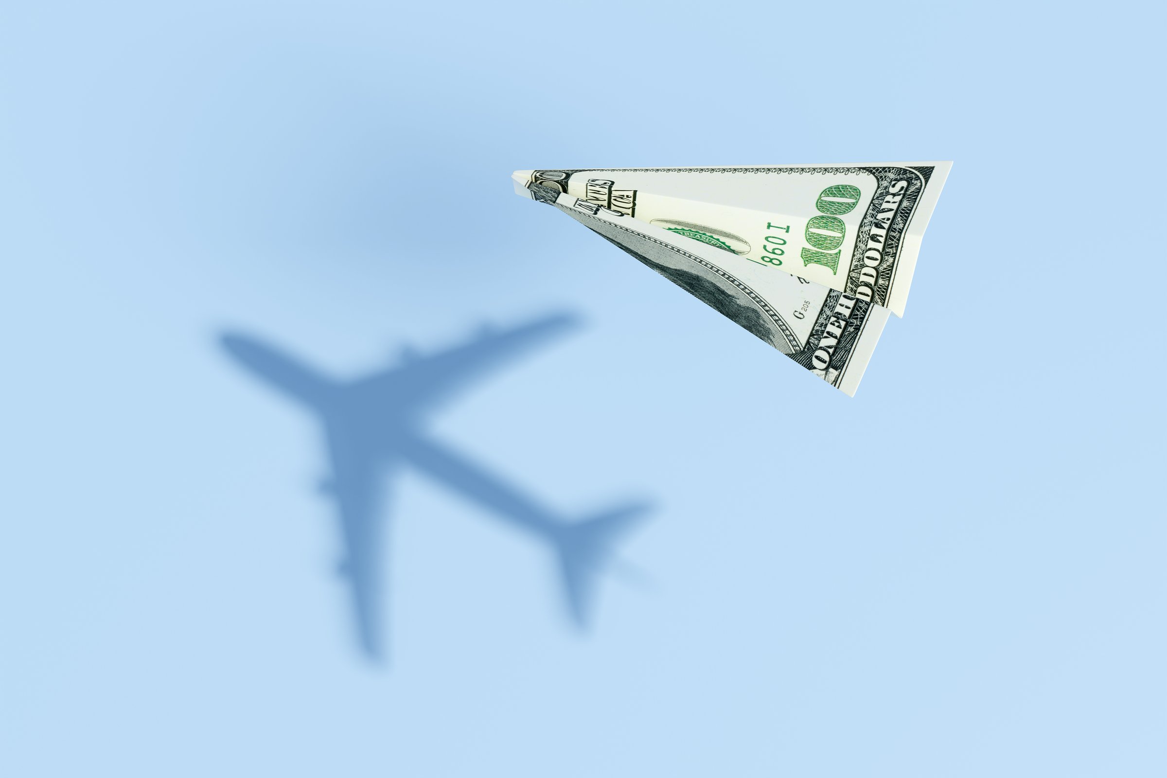 Paper plane made of a US 100 dollar bill flying over a blue sea background and projecting a realistic commercial flight shadow. Illustration of the concept of travel budget and air ticket prices