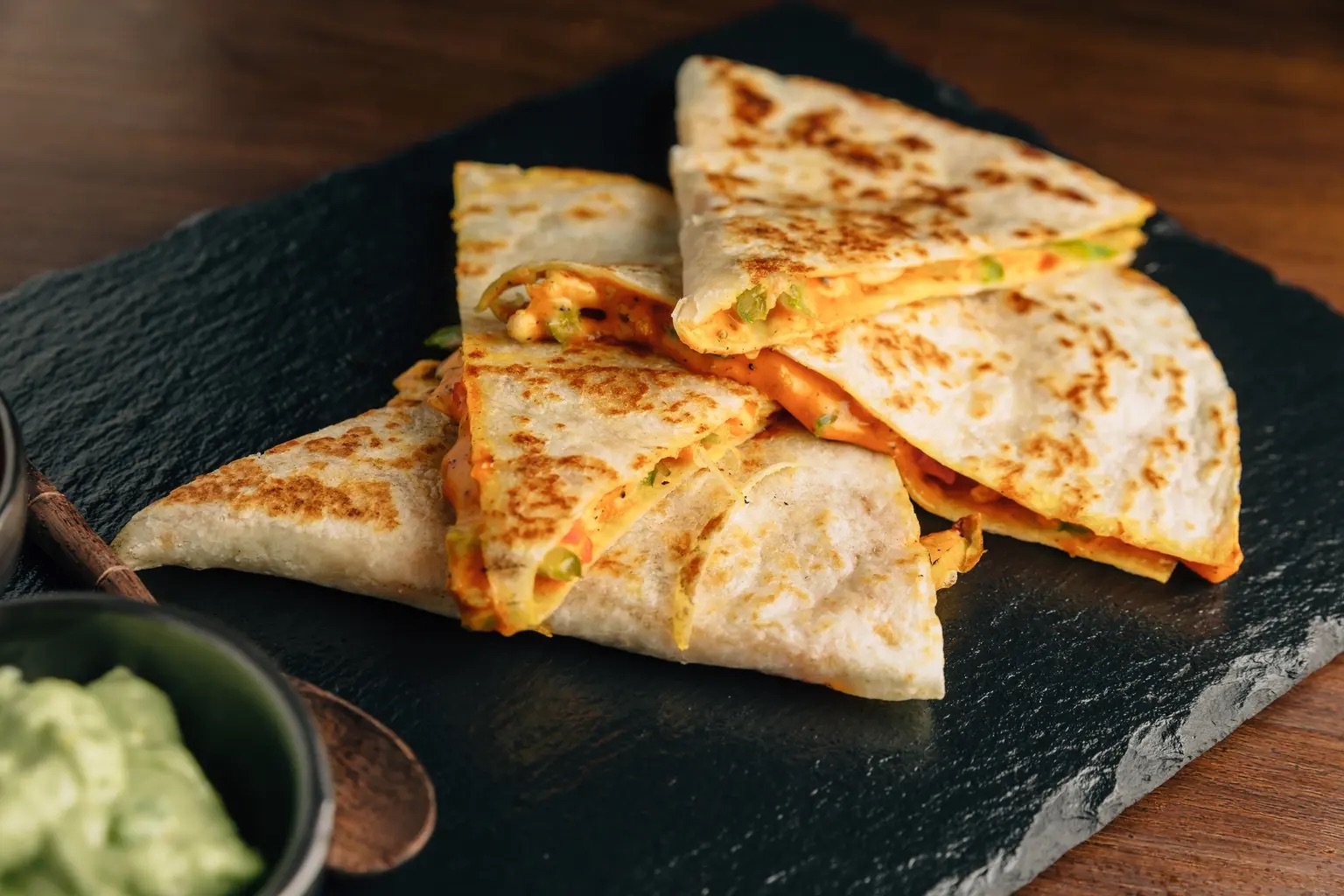 Grilled quesadilla slices with melted cheese and vegetables on a slate board, accompanied by a bowl of guacamole.