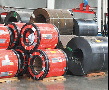 Stacks of large metal coils wrapped in protective red covers, stored on wooden pallets in an industrial setting.