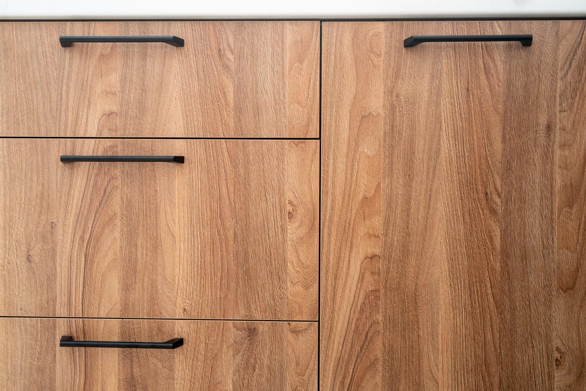 Smooth wooden cabinet with minimalist black handles in a contemporary kitchen. Wooden furniture with closed doors, drawers and natural oak on facade in living room. Front view of cupboard at home