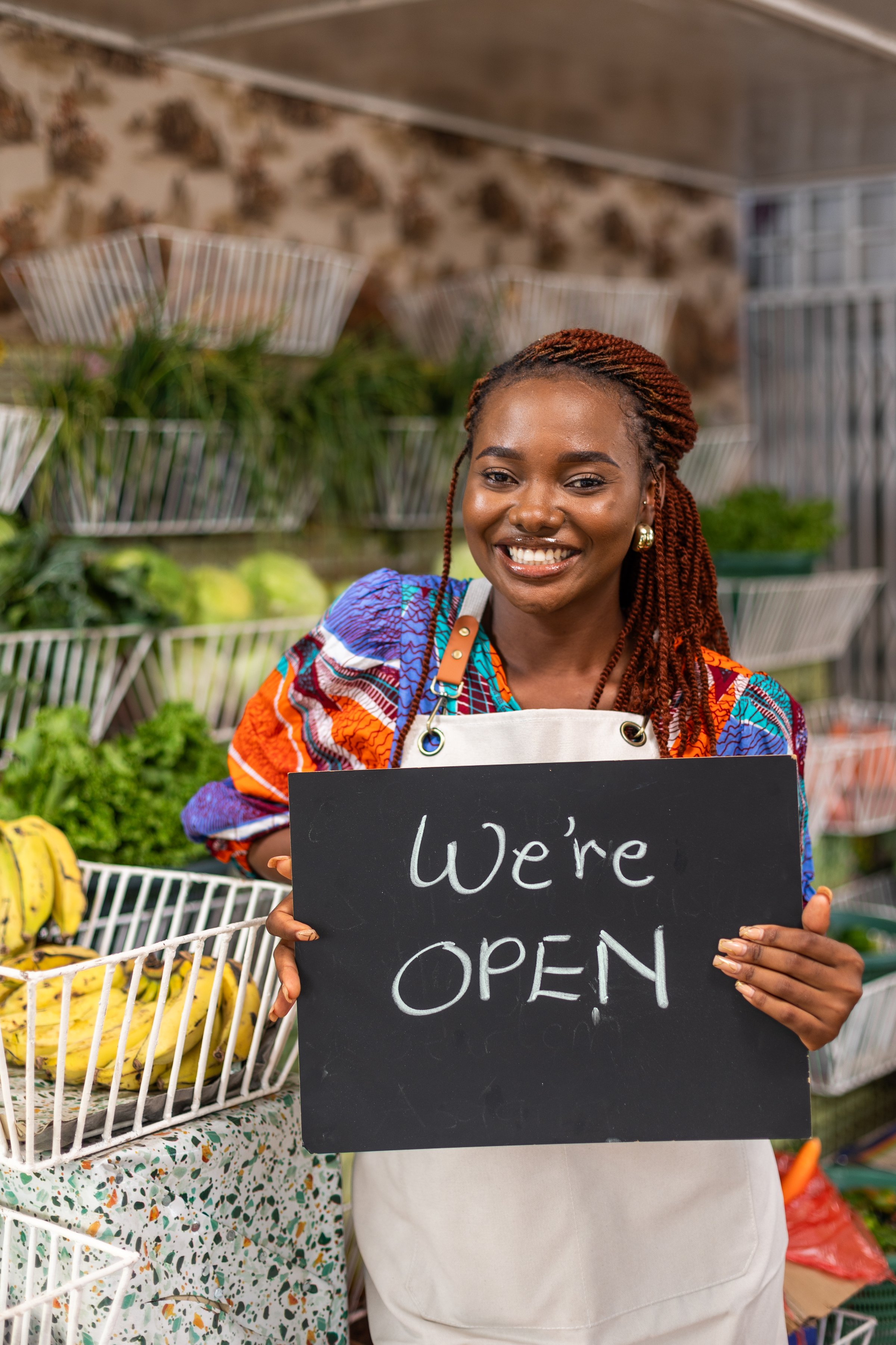 Successful Black female African entrepreneur holding "we are open" sign in front of her grocery shop where she sells healthy fresh fruits and vegetables, in a local market.