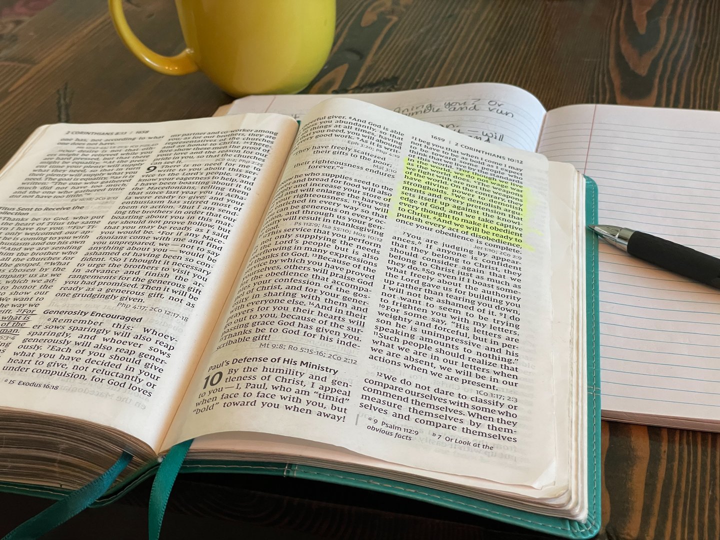 Bible laying open with teal colored book marks. Passage highlighted in yellow. Yellow coffee mug, notebook and pen in background