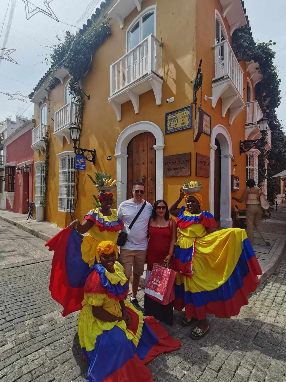 Tourists pose with women in colorful traditional dresses in front of a vibrant building on a cobblestone street.