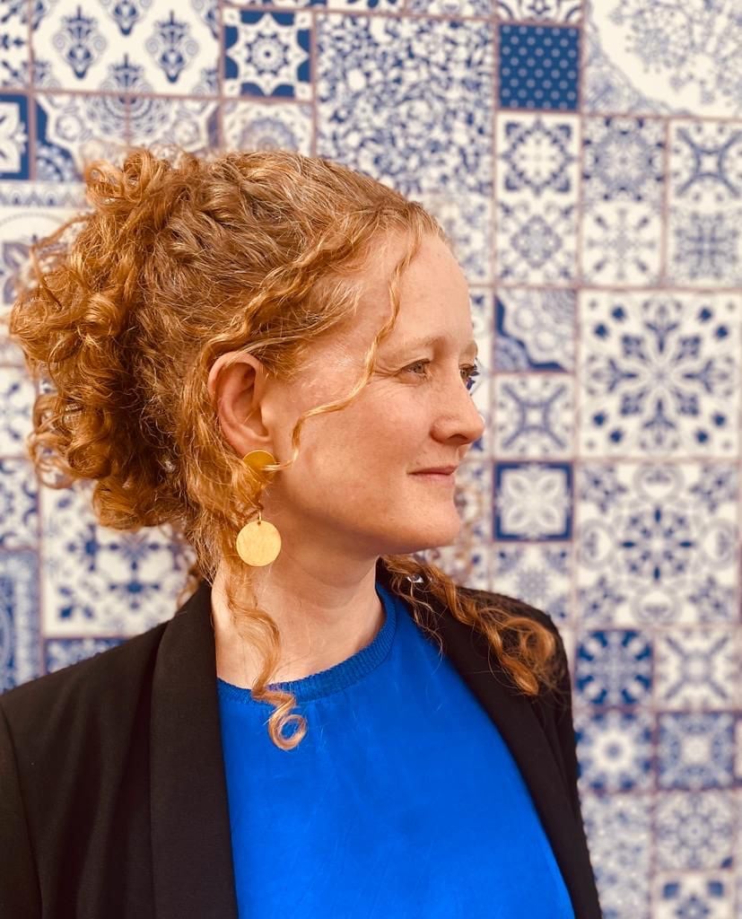 Woman with curly hair, gold earrings, and blue top against a tiled wall with intricate blue and white patterns.