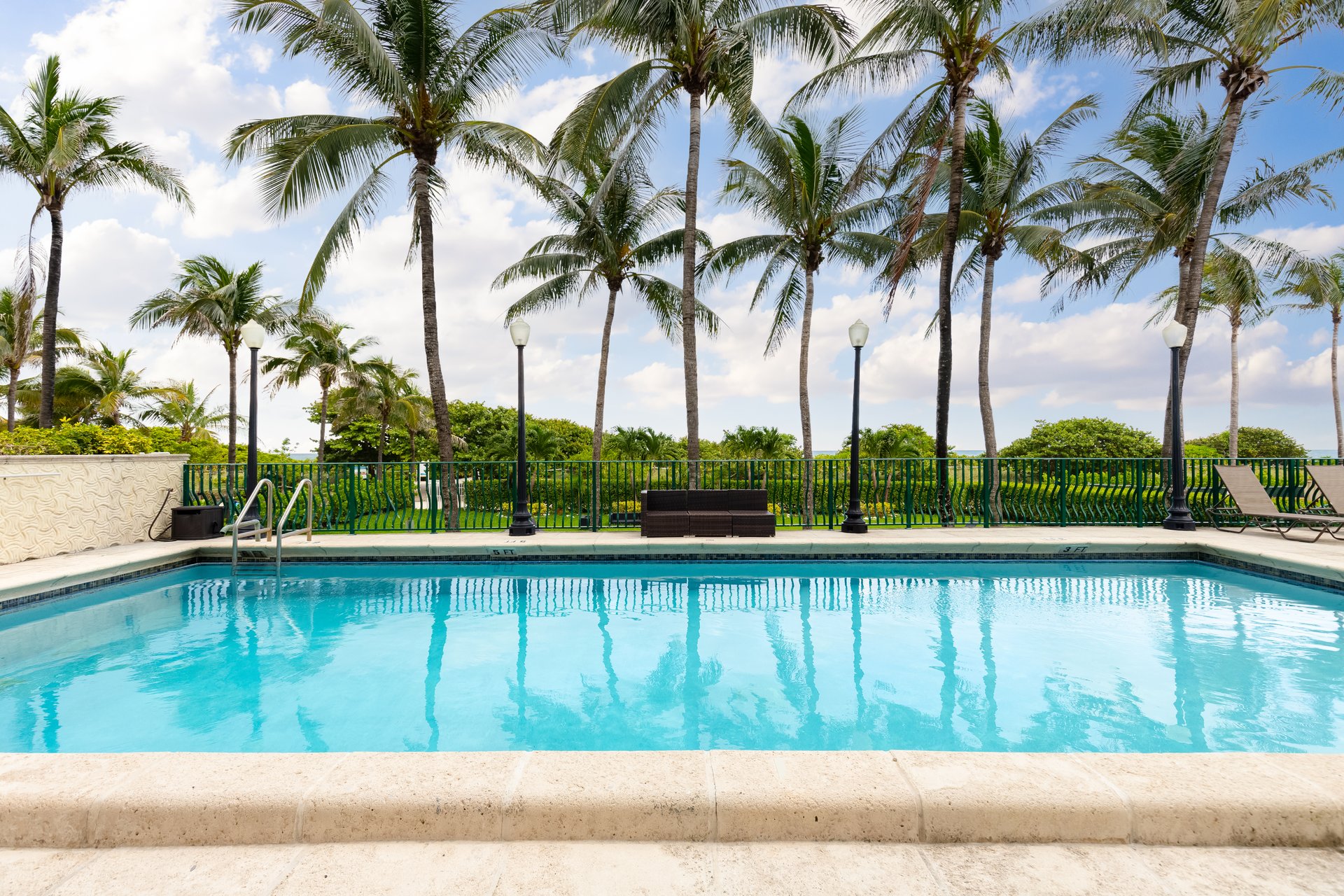 Pool with tropical landscape, surrounded by palm trees, green railing fence with light poles, sun loungers and outdoor furniture, blue sky