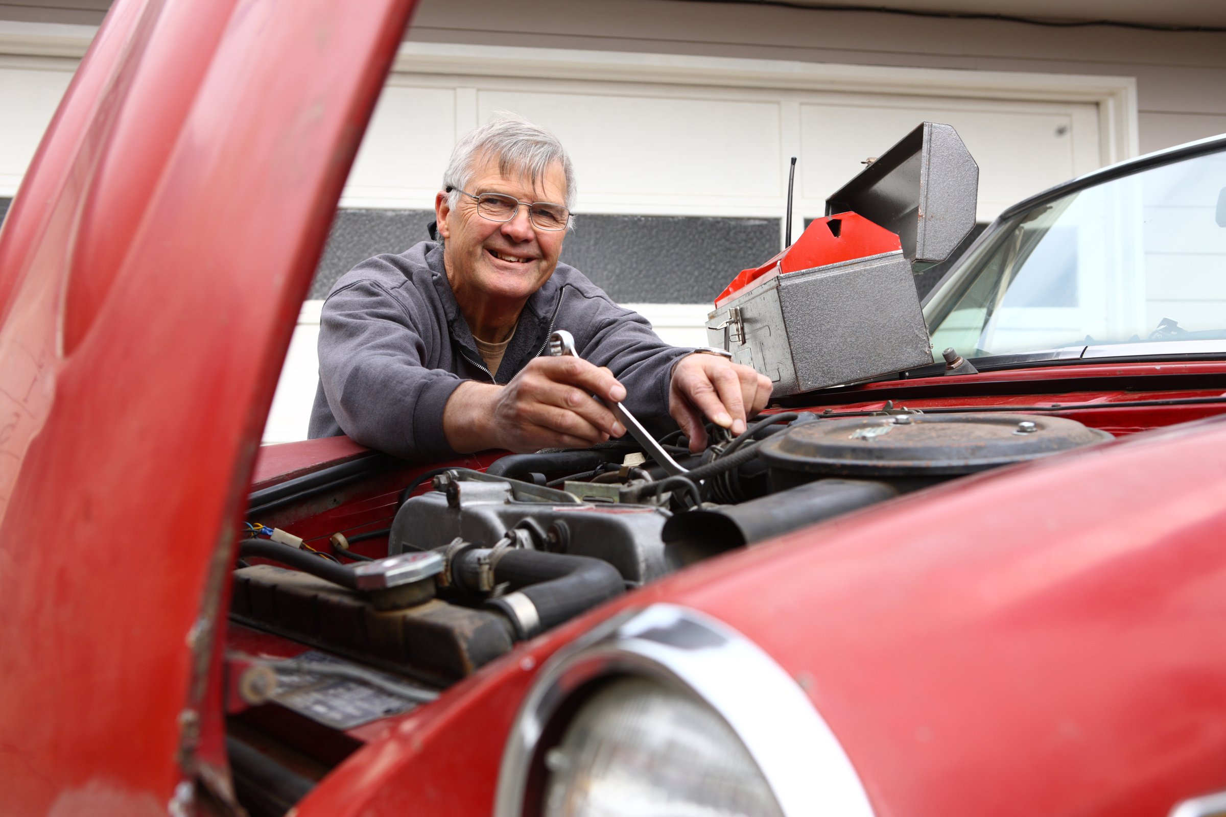 Elderly man smiling while fixing a car engine with a wrench, tool box nearby. Red car hood is open.