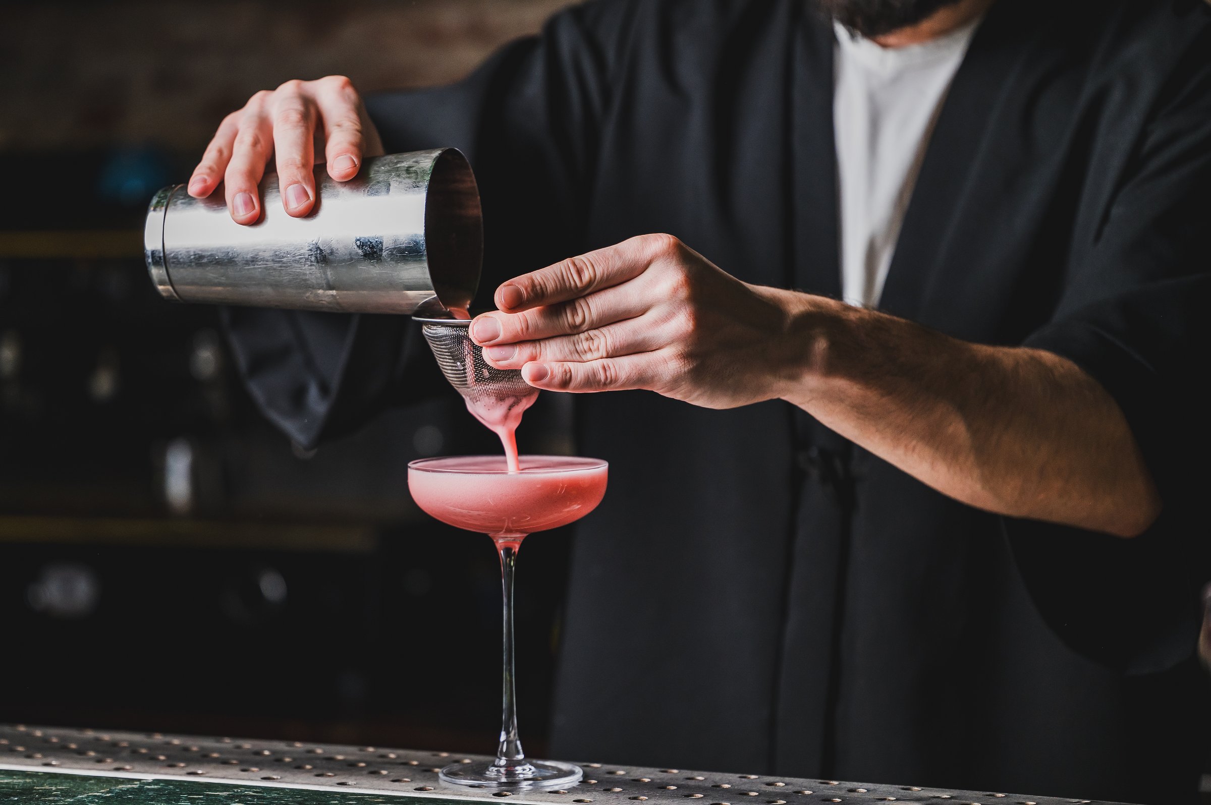 A bartender skillfully pours a vibrant pink cocktail from a shaker through a strainer into a glass. The image highlights precision, craftsmanship, and the art of mixology in a bar setting