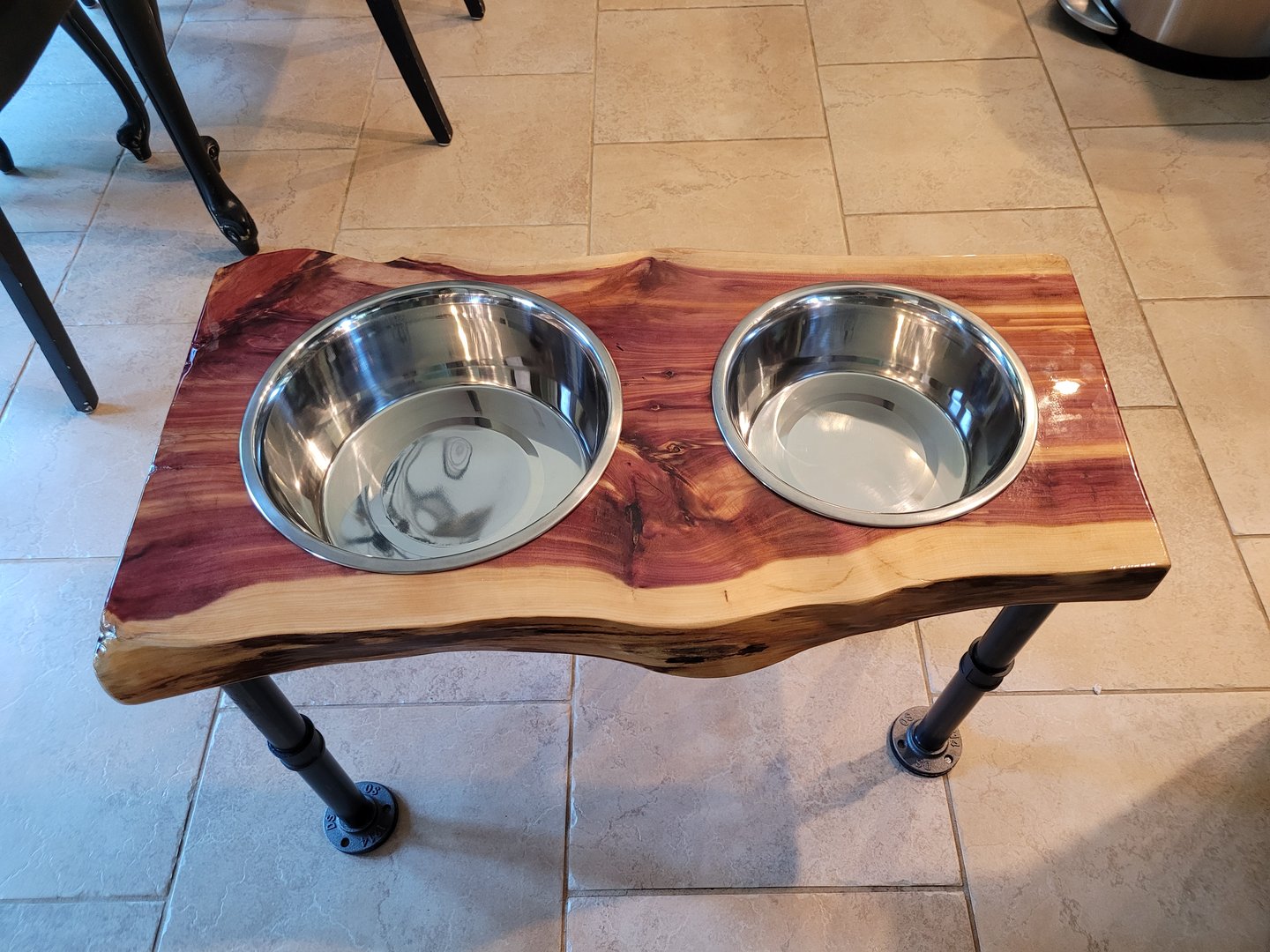 Rustic elevated pet feeder with two stainless steel bowls on a wooden stand with black metal legs, set on a tile floor.