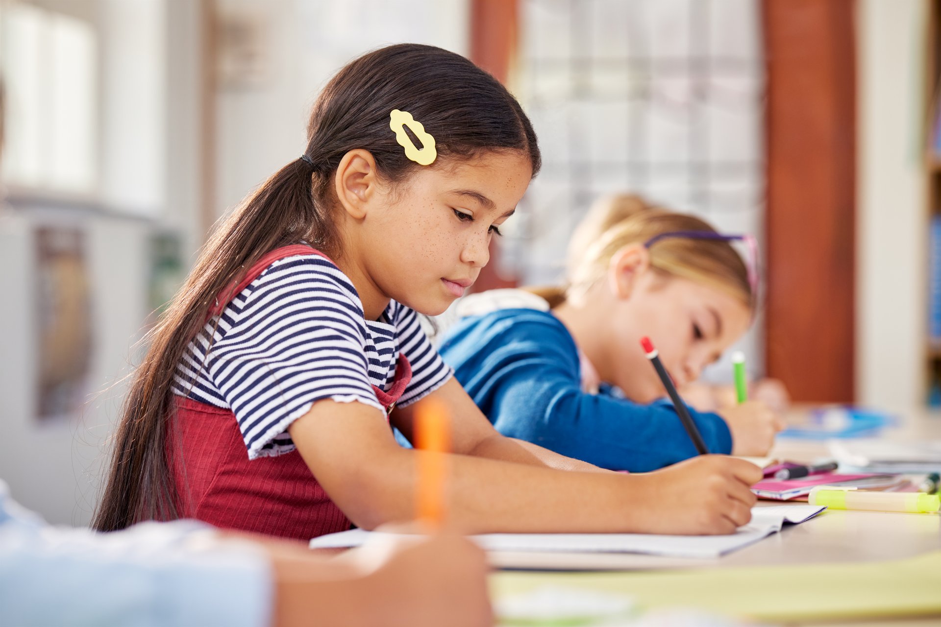 Cute little school girl with the two pigtails writing in book with classmates in background. Little pupil writing at desk in classroom at the elementary school. Student girl study doing test in primary school.