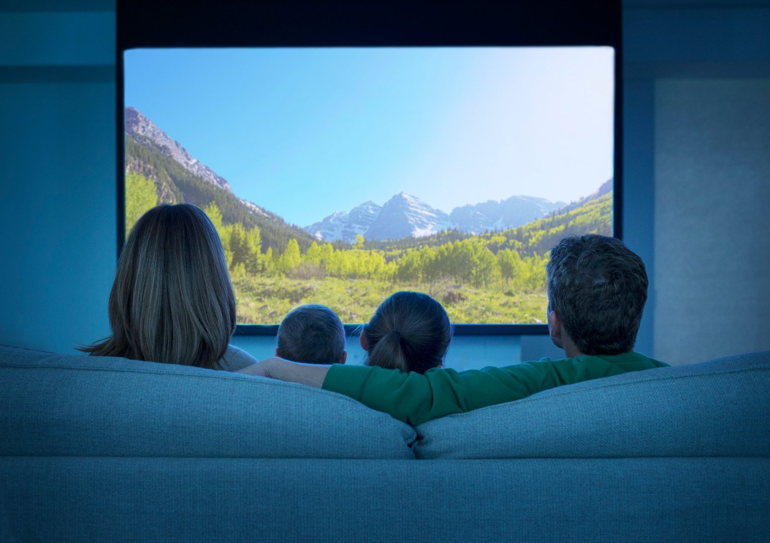Family of four watching nature scenery on a large TV, seated on a couch, with a view of mountains and greenery.