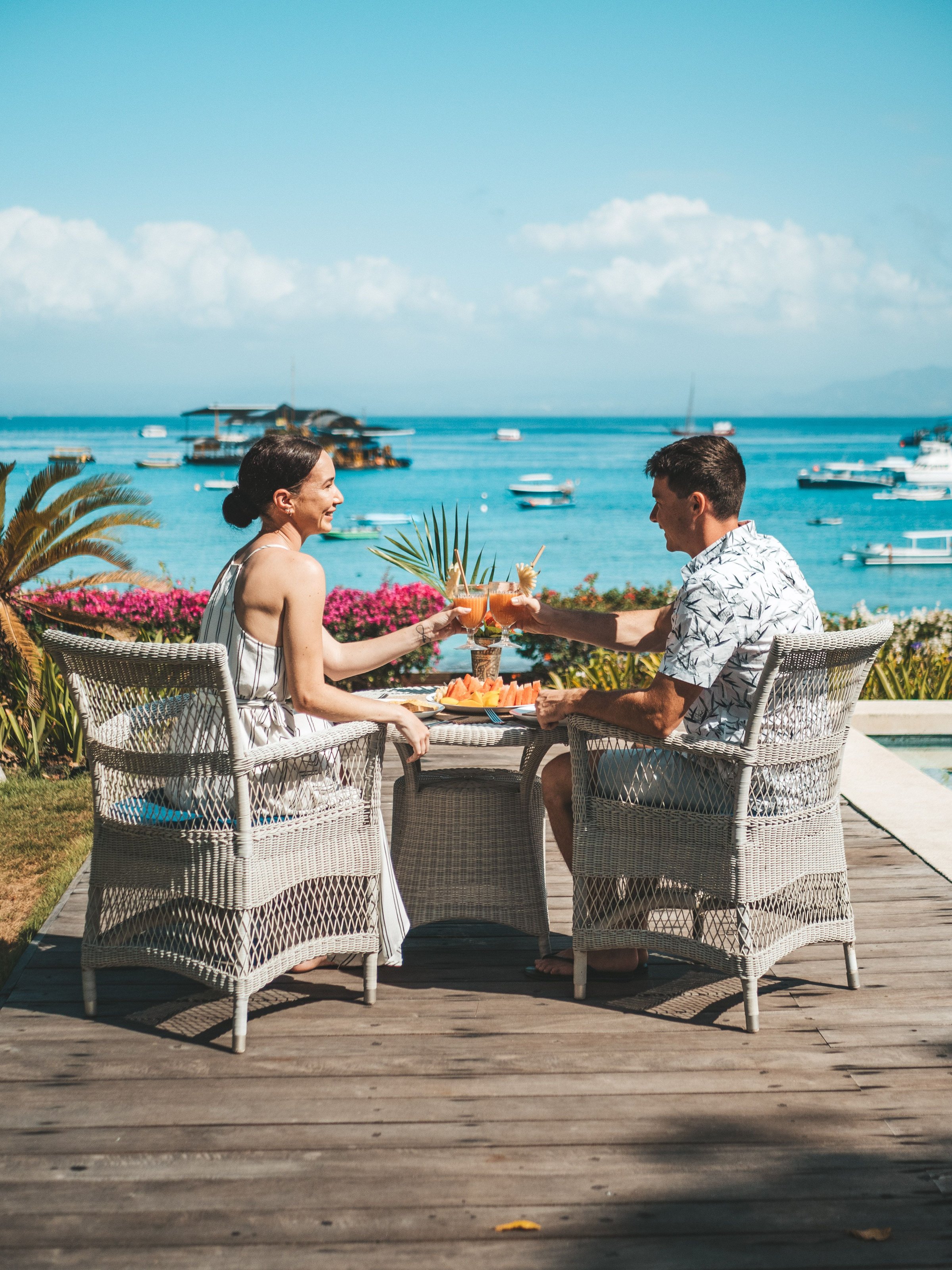 A couple enjoying drinks and a meal by the seaside with boats in the background on a sunny day.