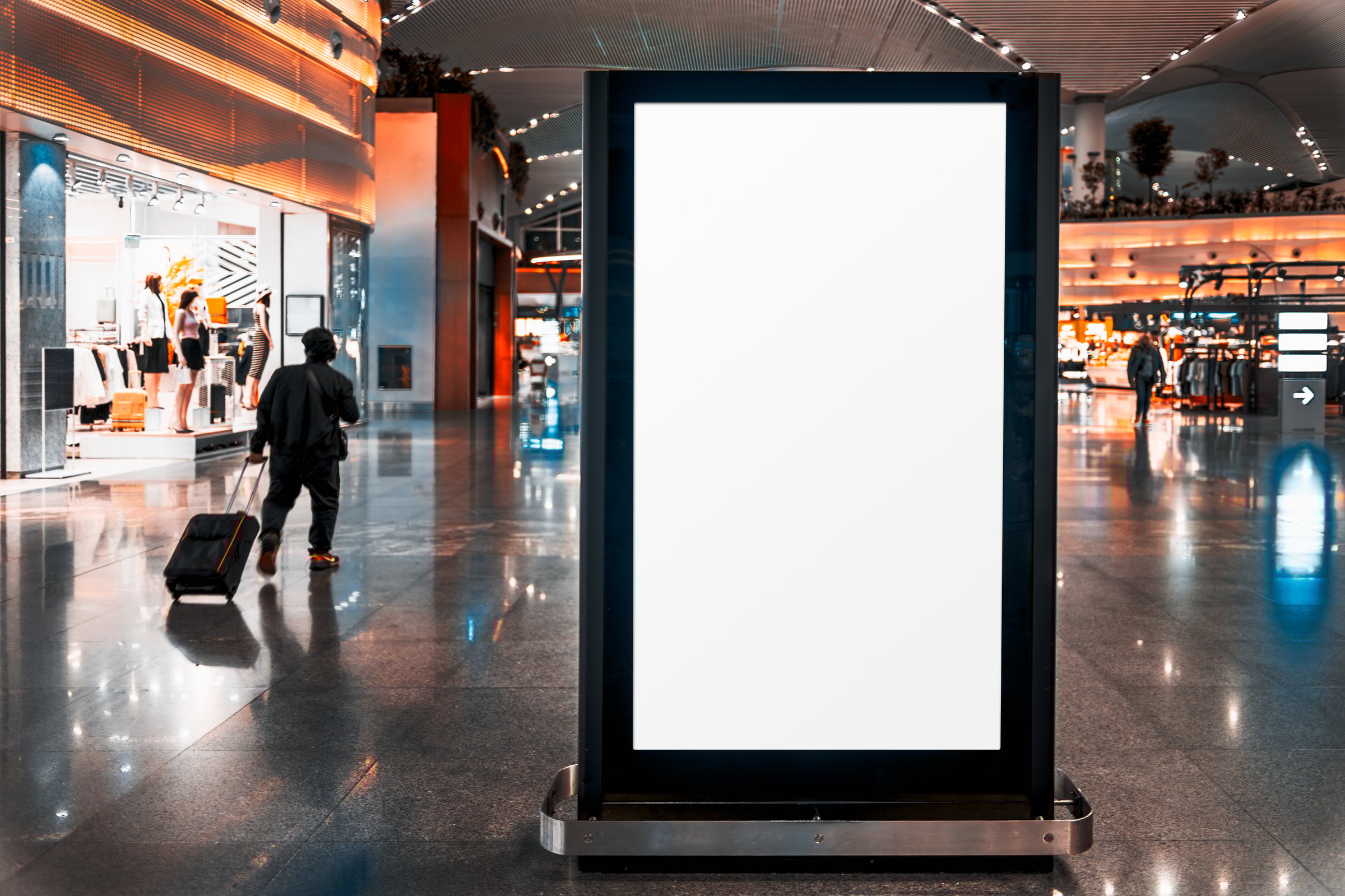A modern airport terminal with a large digital advertising screen mockup - a blank white space in the foreground. A silhouette of a traveler pulling a suitcase walks past a fashion retail store