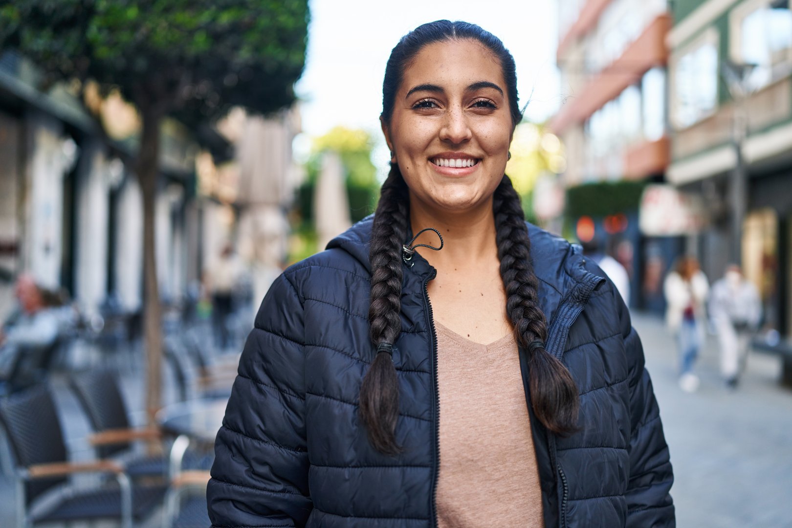 Smiling woman with long braided hair wearing a dark jacket stands on a busy pedestrian street lined with buildings and trees.
