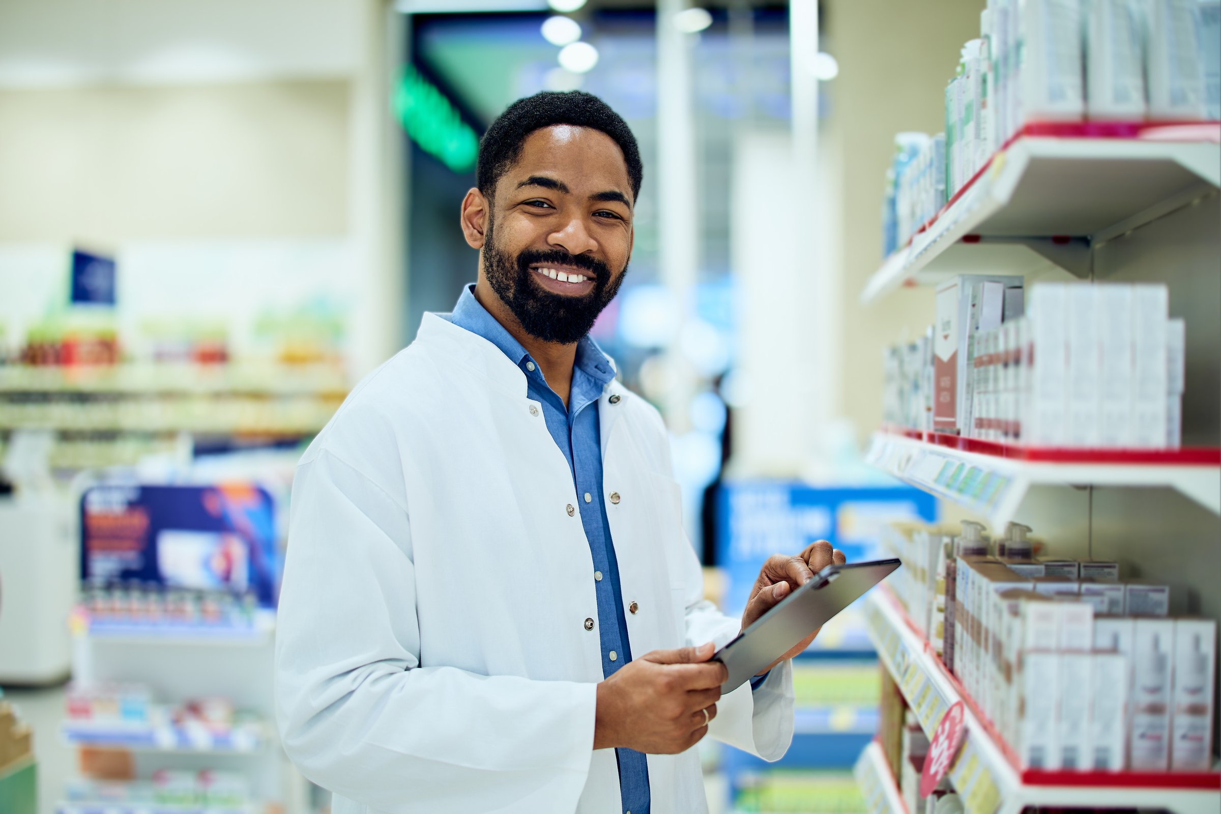 Friendly pharmacist standing in a pharmacy aisle, holding a tablet and smiling.