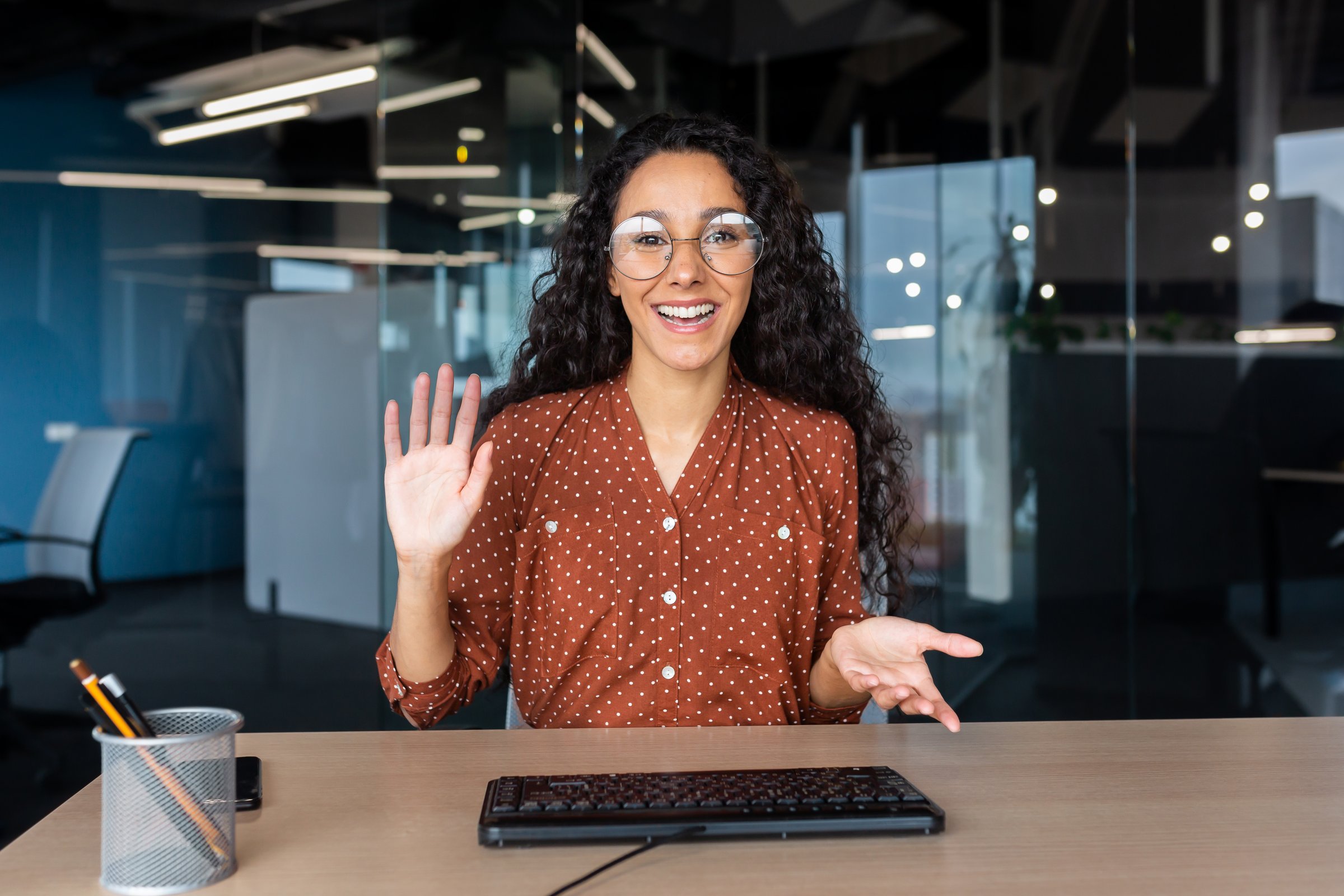 Online customer support video call, Latin American woman looking at web camera smiling and consulting customers, helpline worker working inside office.