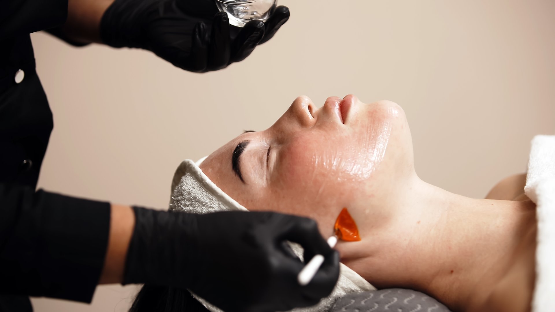 Close-up of a cosmetologist applying a cosmetic peeling mask to a client's face. The scene emphasizes the professional approach to skin care and the relaxing spa experience.