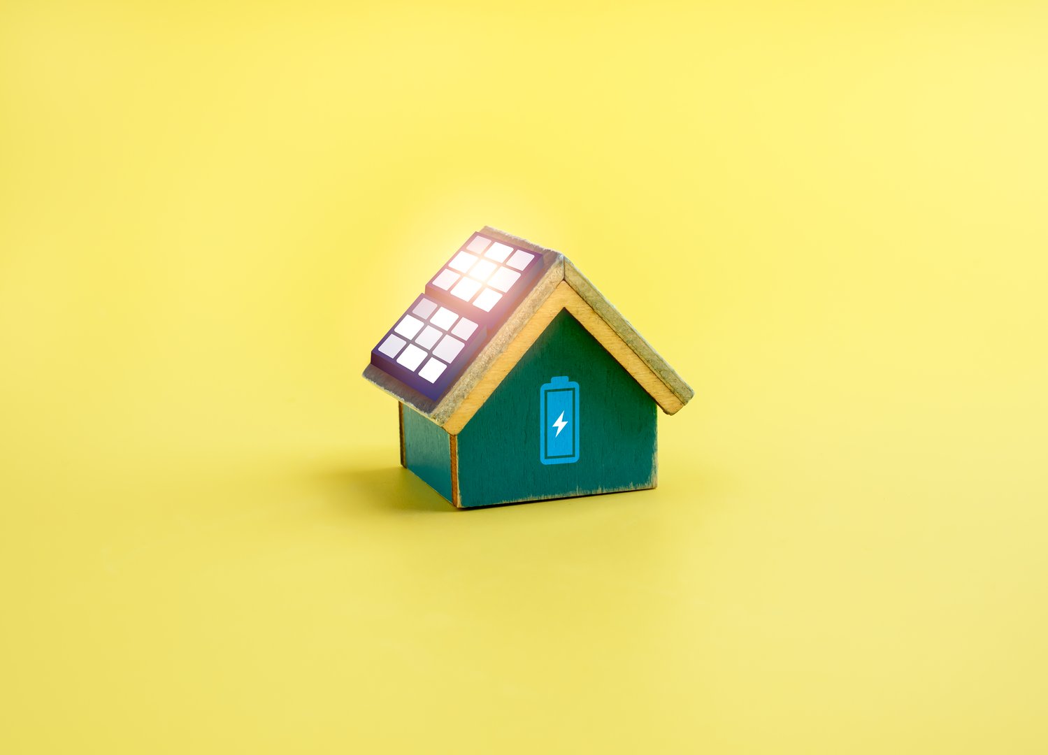 Miniature green house model with solar panels and battery icon on yellow background, symbolizing energy-efficient home, clean energy, sustainable living, and renewable power solutions.