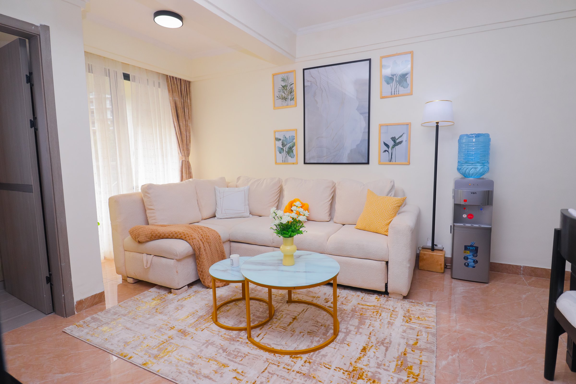 Cozy living room with a beige sectional sofa, coffee table, floral art, floor lamp, and water dispenser.