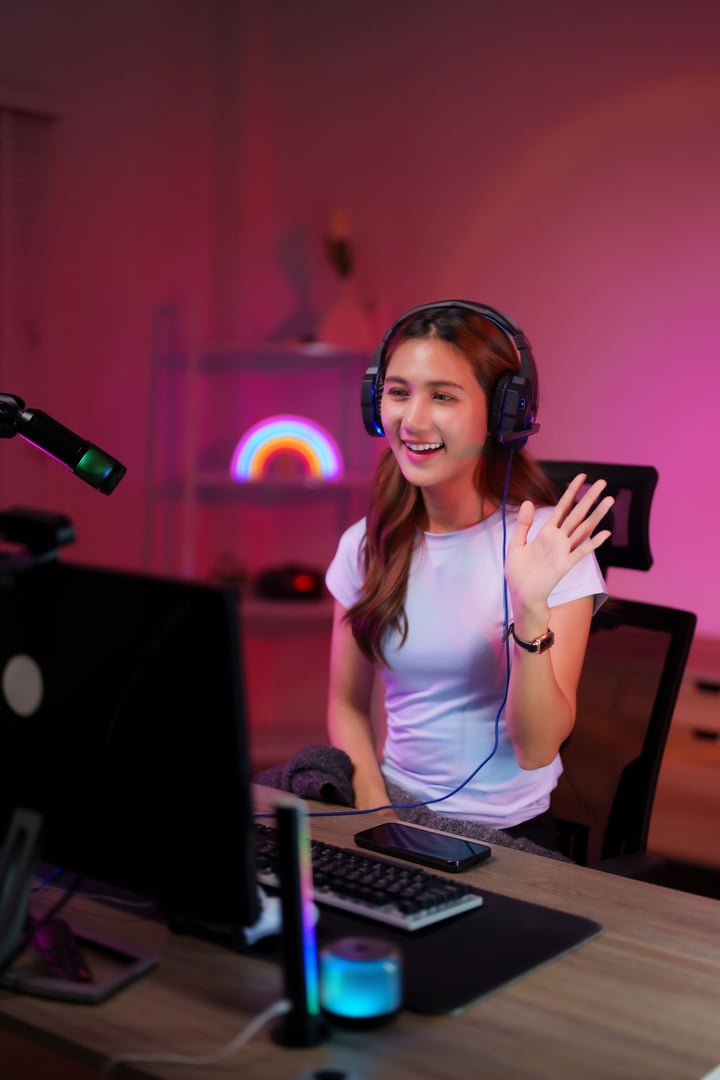 Happy Asian streamer broadcasts online game session from colorful room wearing headset greeting viewers during technology broadcast